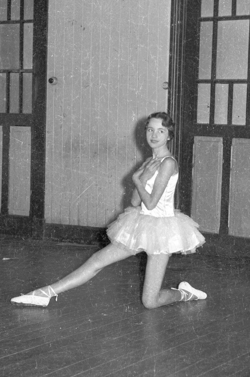 Retrato de plano entero de una mujer joven caracterizada como bailarina de ballet. está posando hincada y de Medio perfil. Se encuentra en en el interior de un lugar no identificado