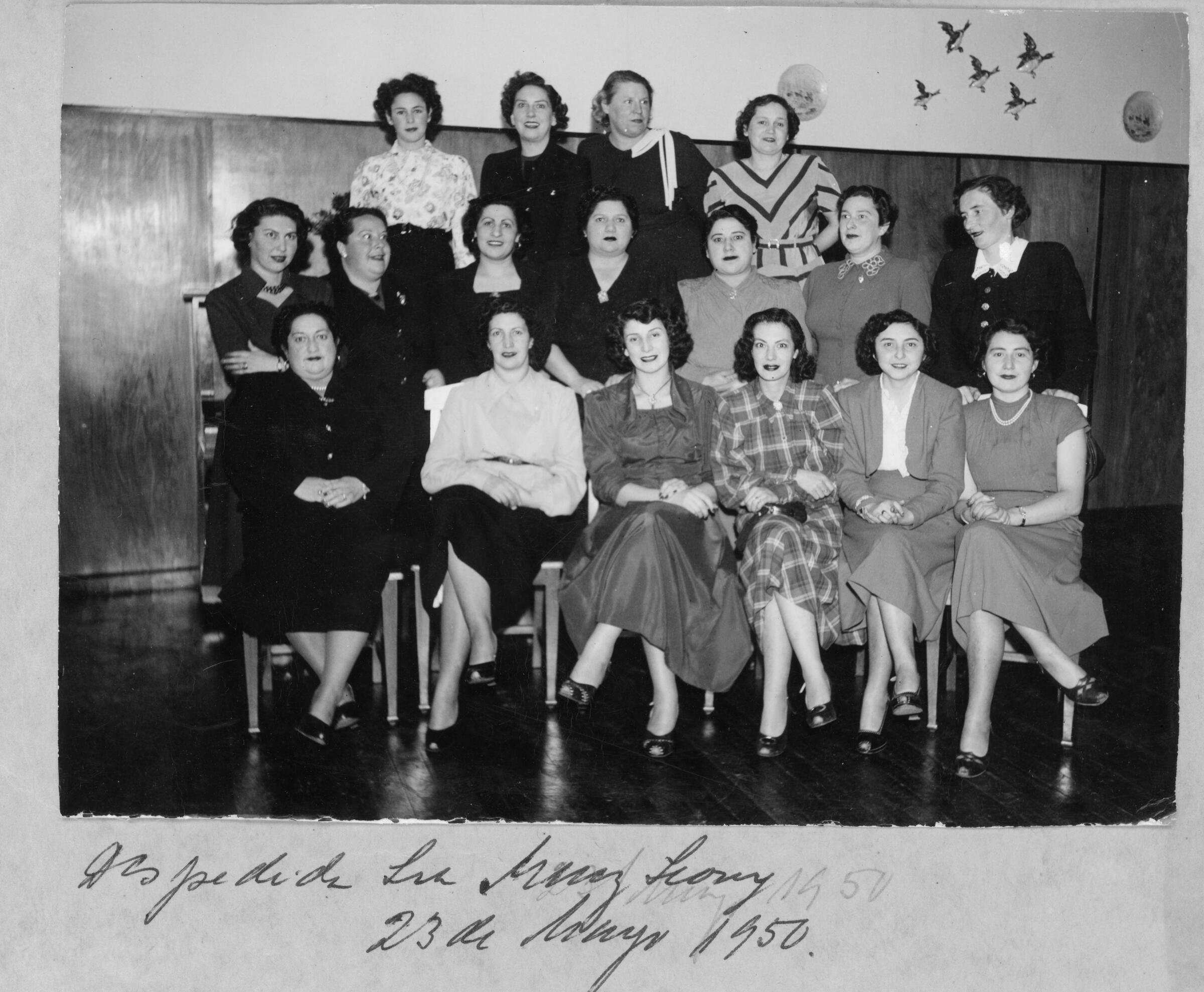 Retrato grupal de plano entero de Mujeres formadas en tres hileras mientras posan. La tercera mujer de izquierda a derecha, que se encuentra de pie en la segunda hilera es Olga Boettcher. Están en el interior de una infraestructura no identificada, durante una despedida realizada para una de las mujeres.