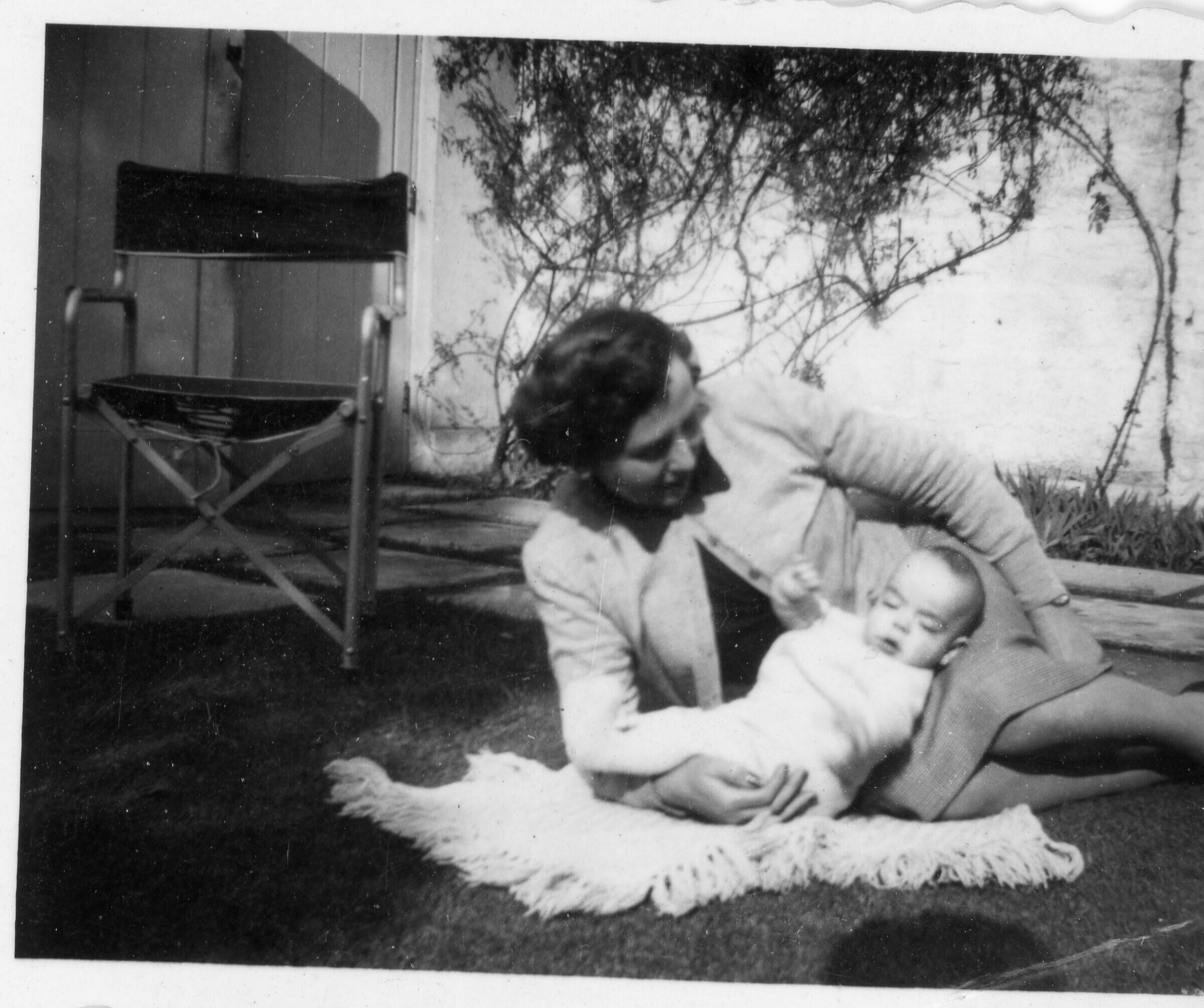 Retrato de plano entero de una mujer recostada en el pasto, mirando a un bebé que está de espaldas sobre una manta, apoyando su cabeza sobre sus piernas. Se encuentran en el patio de una casa