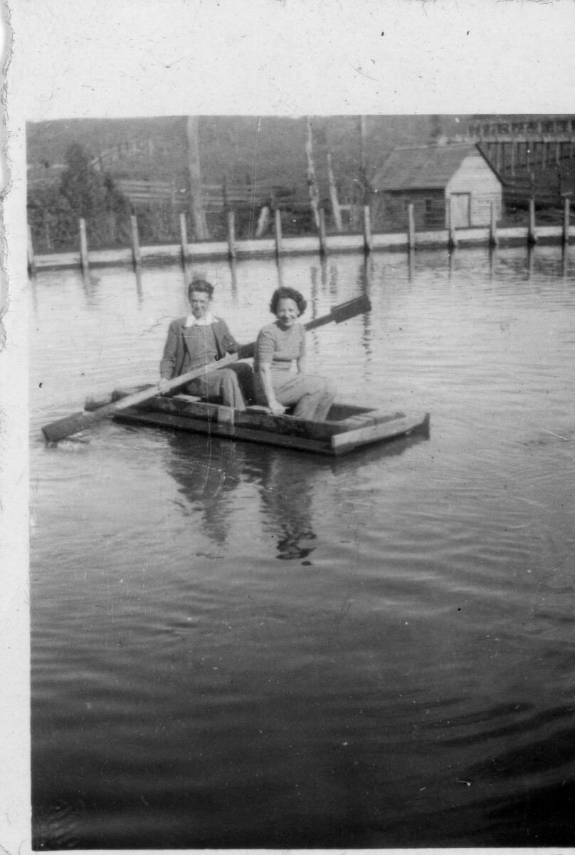 Retrato de plano entero de dos mujeres posando en el interior de un bote. Una de ellas está sosteniendo el remo. El lugar está cercado y en el fondo se observa una casa de madera. Lugar no identificado.