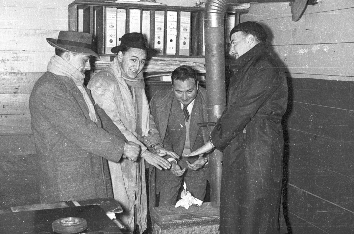 -Plano entero de cuatro hombres calentando sus manos alrededor de una  estufa a leña. detrás de ellos, junto a la pared, hay un estante con una hilera de archiveros ubicados en la repisa superior, los cuales tienen escrito en el lomo un texto ilegible, a excepción de uno que dice -- Libre tratado de vinos, licores y tabacos - 1953--. Están en el interior de la Caja Nacional de Ahorros-