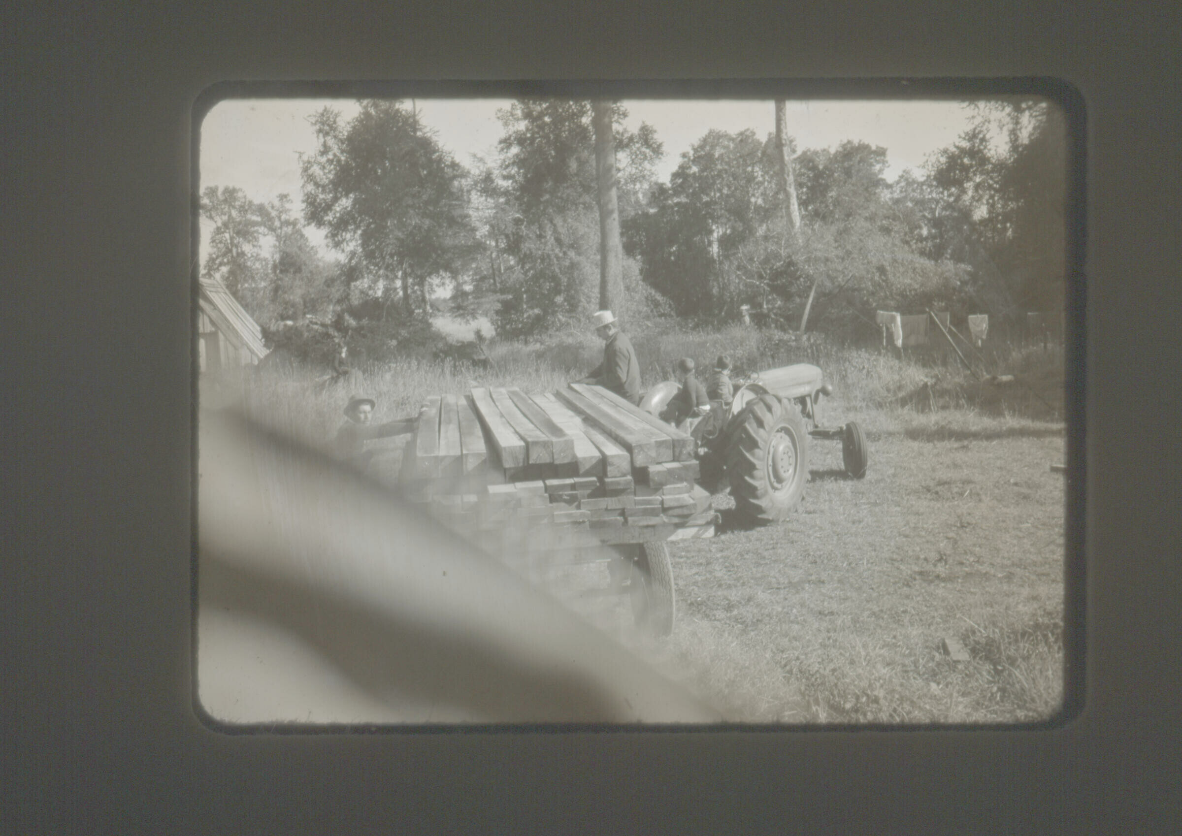 Retrato de plano entero de dos niños de temprana edad sobre un tractor que lleva una carreta con tablas de maderas, donde trabajan dos hombres