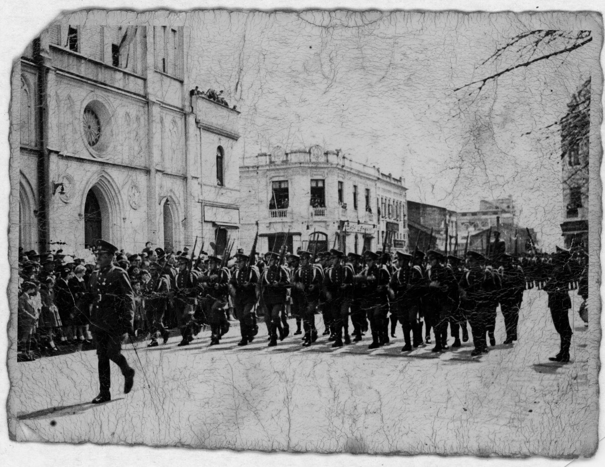 plano general de un grupo uniformados, formados en hileras mientras desfilan con sus fusiles por la calle O'Higgins de Valdivia. En un costado, un grupo de Niñas, Niños, Mujeres y Hombres los observan desde la vereda.