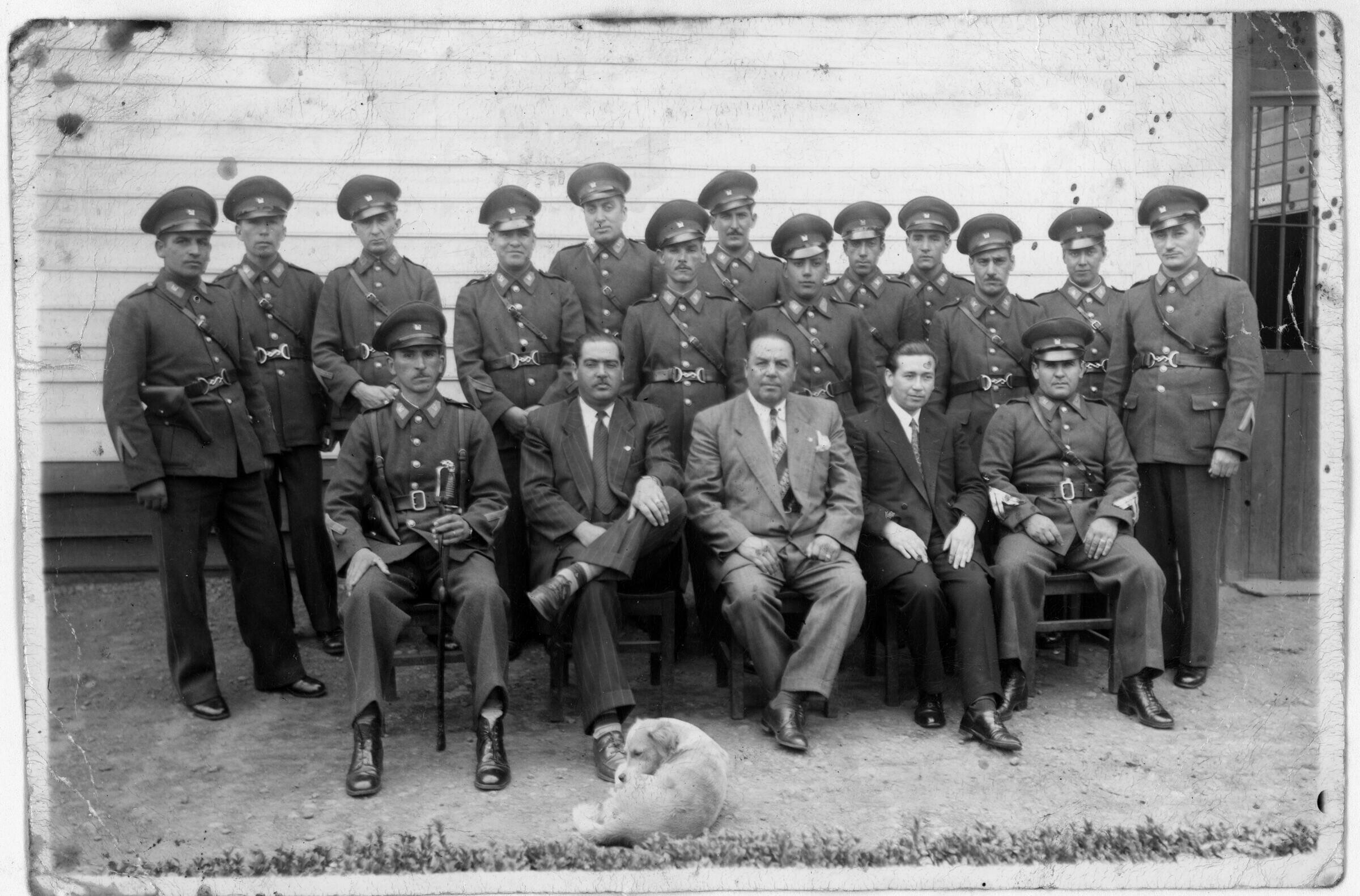 Retrato grupal de plano entero de uniformados de carabineros posando junto a tres hombres vestidos de Traje, los cuales se encuentran sentados en primera fila mientras que el resto están parados por detrás. En el frente, hay un perro durmiendo en el piso. Están ubicados junto a una pared en el interior de la tercera comisaría