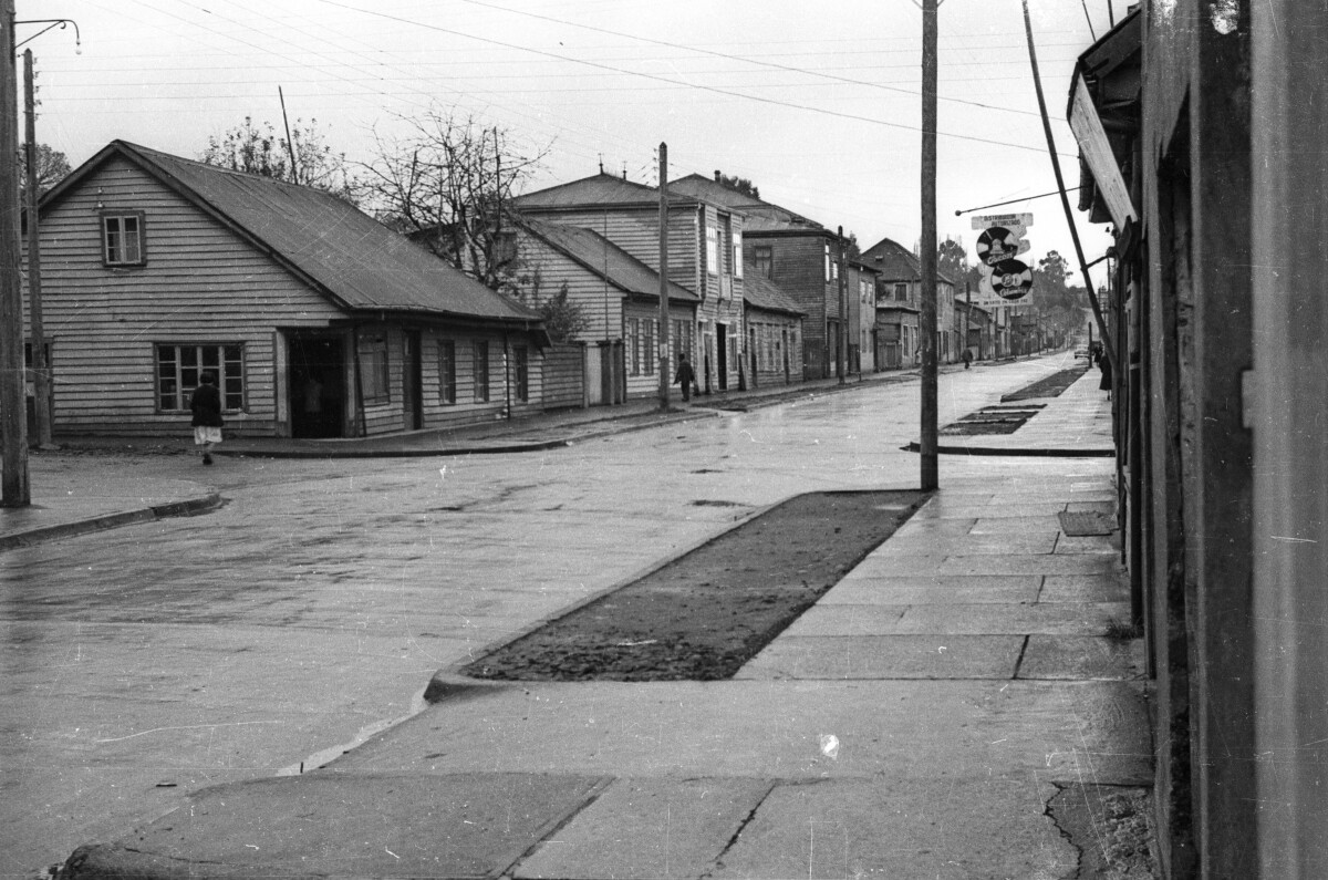 Vista general de la intersección de dos calles en las cuales hay casas y tiendas. La fotografía fue tomada durante un día de lluvia.