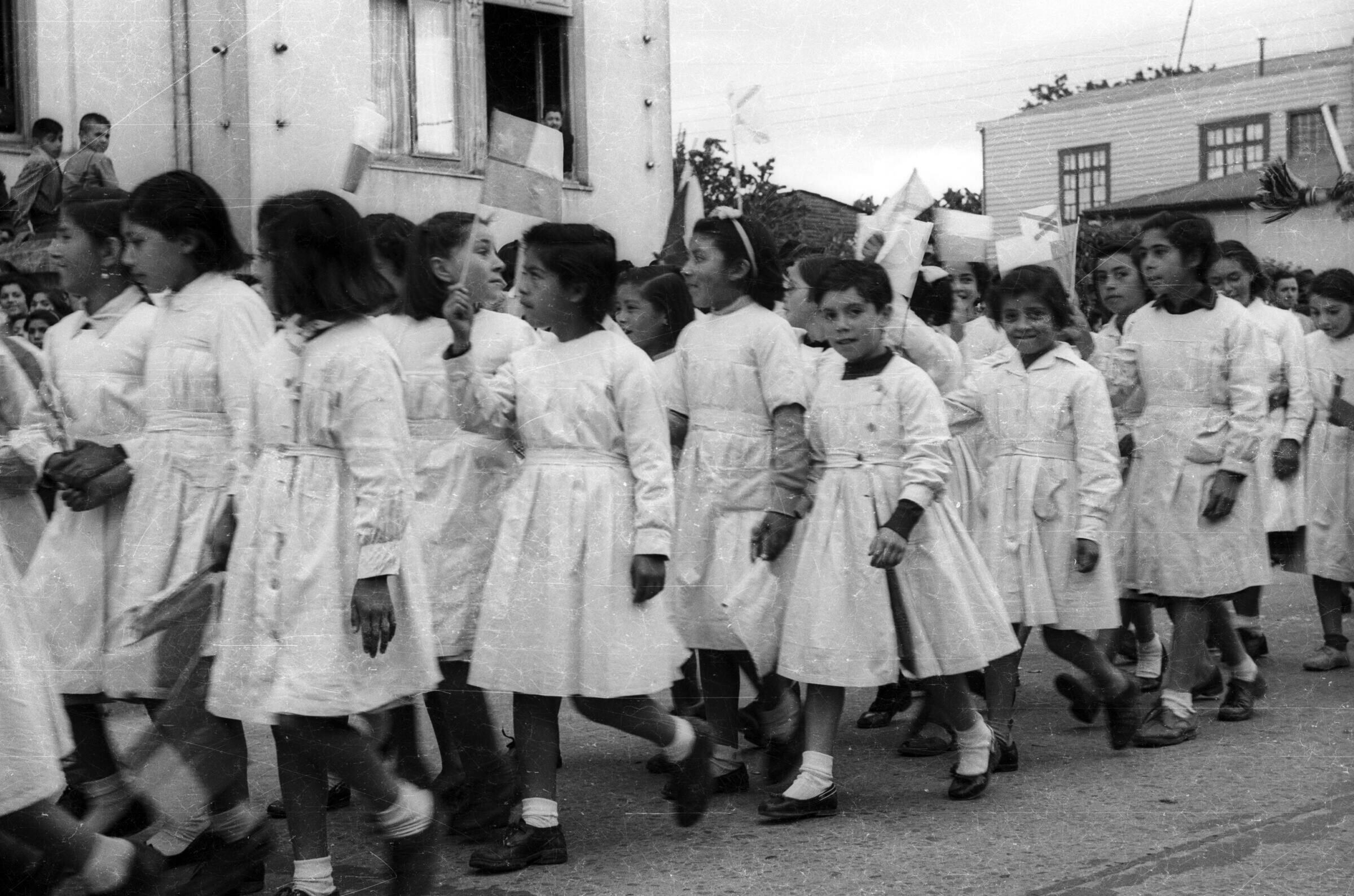 Plano entero de un grupo de niñas con delantal, desfilando formadas en hileras, con una bandera en las manos, por el frontis de la municipalidad