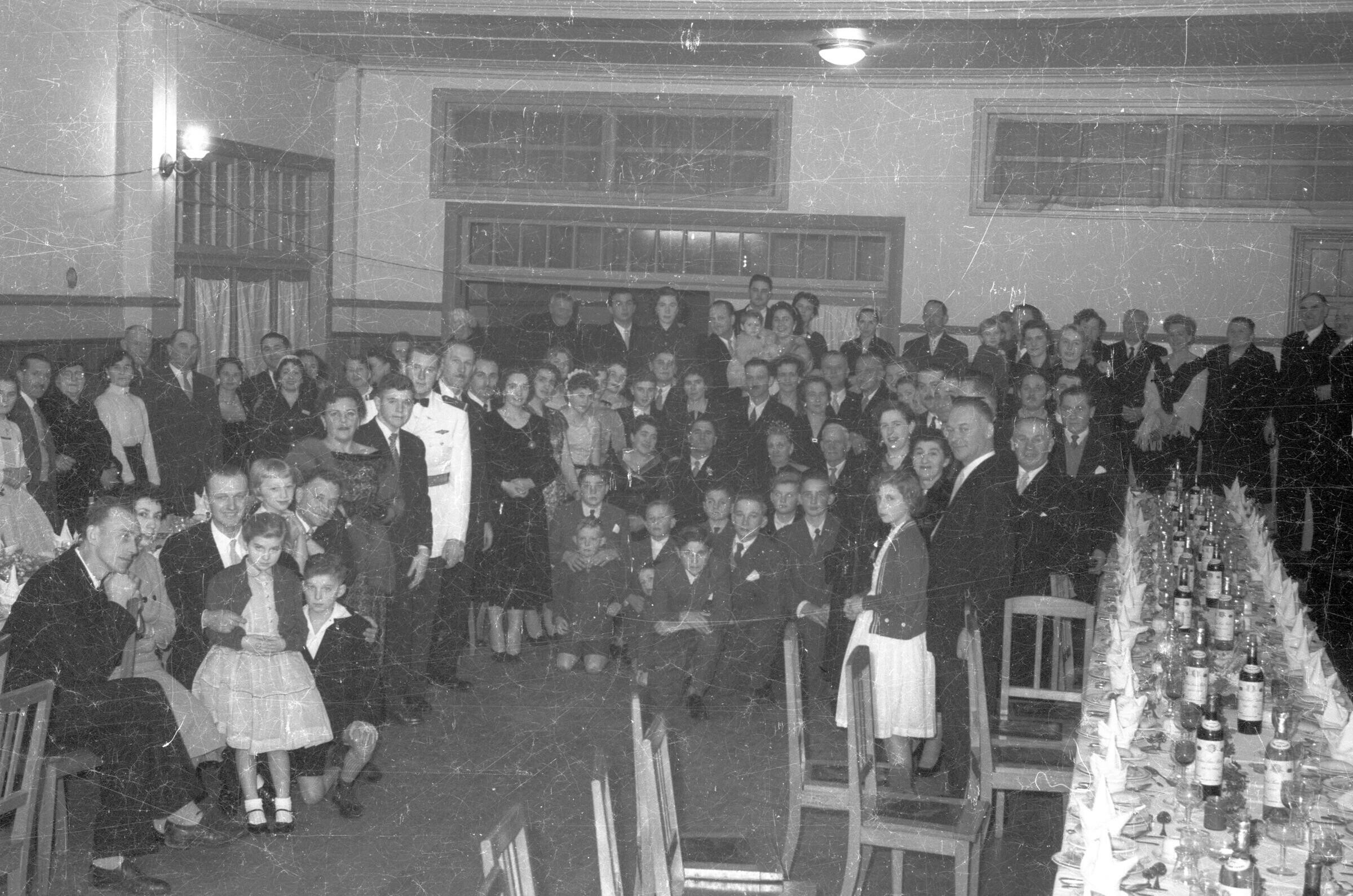 Retrato grupal de plano entero de grupo de Hombres con Trajes, Mujeres con Vestidos, niños y niñas, ubicados todos juntos al final de una sala, donde hay una mesa servida con vinos y vajilla.