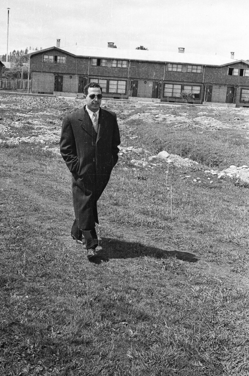 Plano entero de un hombre caminando, va vestido de traje, gafas, lleva las manos en los bolsillos de su largo abrigo, en el fondo hay casas y va caminando sobe el pasto de un terreno abierto
