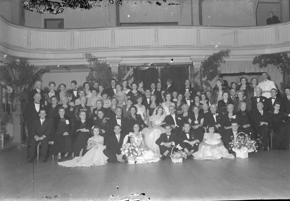 Plano general de un grupo de personas posando para un retrato grupal junto a una pareja de recién casado, sentados al centro del grupo