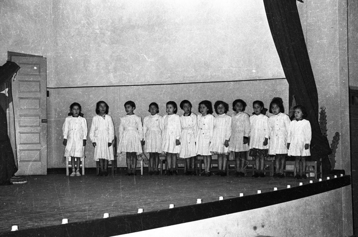 Plano entero de un grupo de Niñas con Vestido, paradas junto a sus sillas sobre el escenario mientras se lleva a cabo una presentación artística en el gimnasio de la escuela de Niñas Nº2