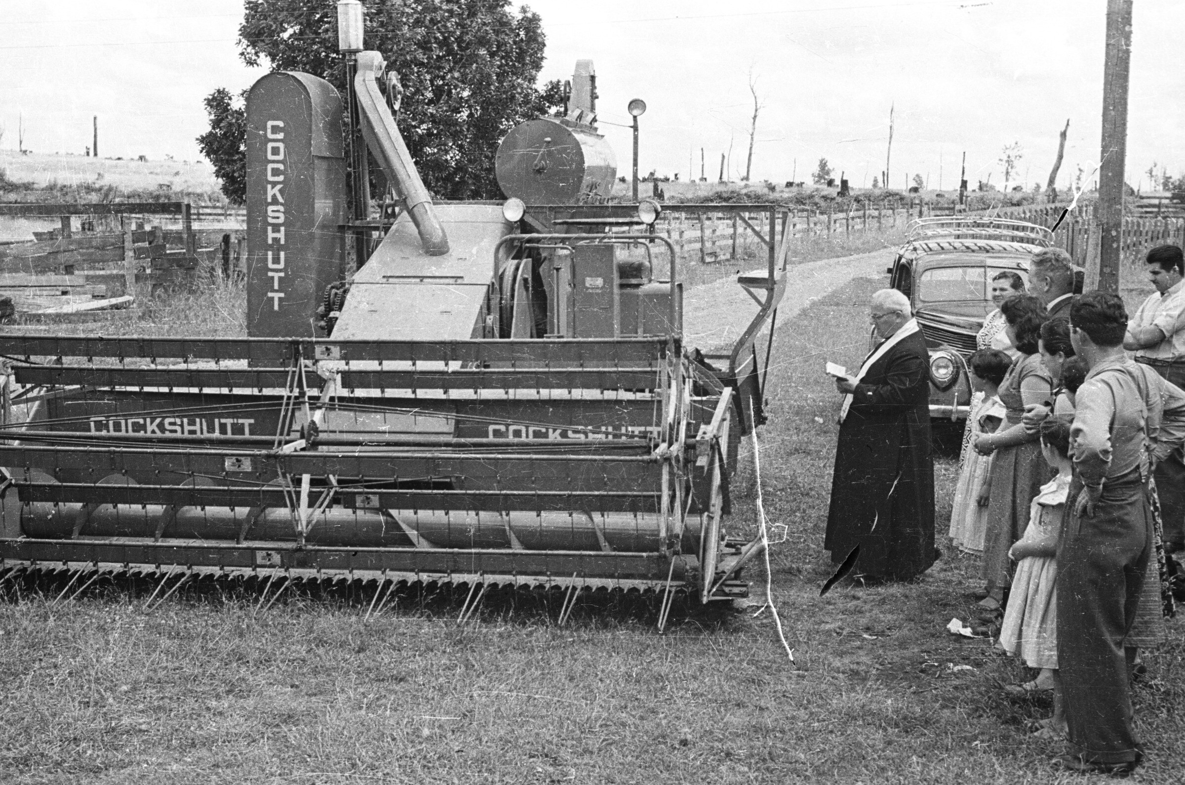 plano general de un sacerdote bendiciendo una maquina cosechadora, marca cockshutt. Lo acompañan un grupo de Hombres, Mujeres y Niñas. La maquina está ubicada al costado de un camino de tierra en una zona rural no identificada