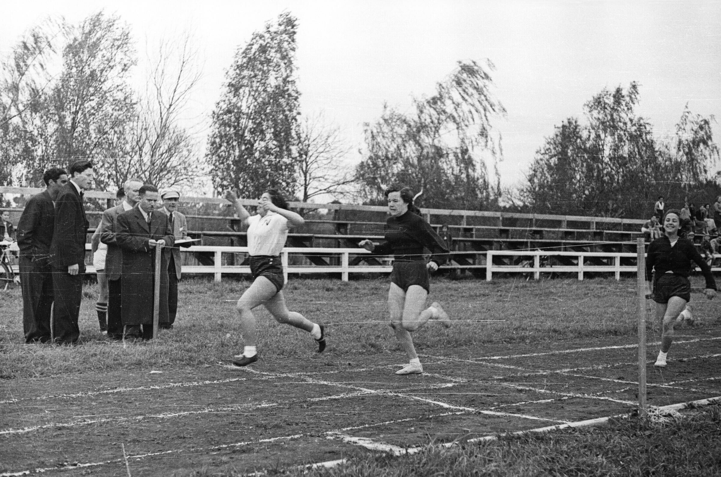 Plano entero de tres mujeres corriendo próximas a llegar a la meta durante una carrera, a su alrededor hay un grupo de hombres que las observa, uno de ellos está anotando en una libreta. La carrera se está llevando a cabo dentro de una jornada de competencias de juegos olímpicos dentro de un recinto abierto