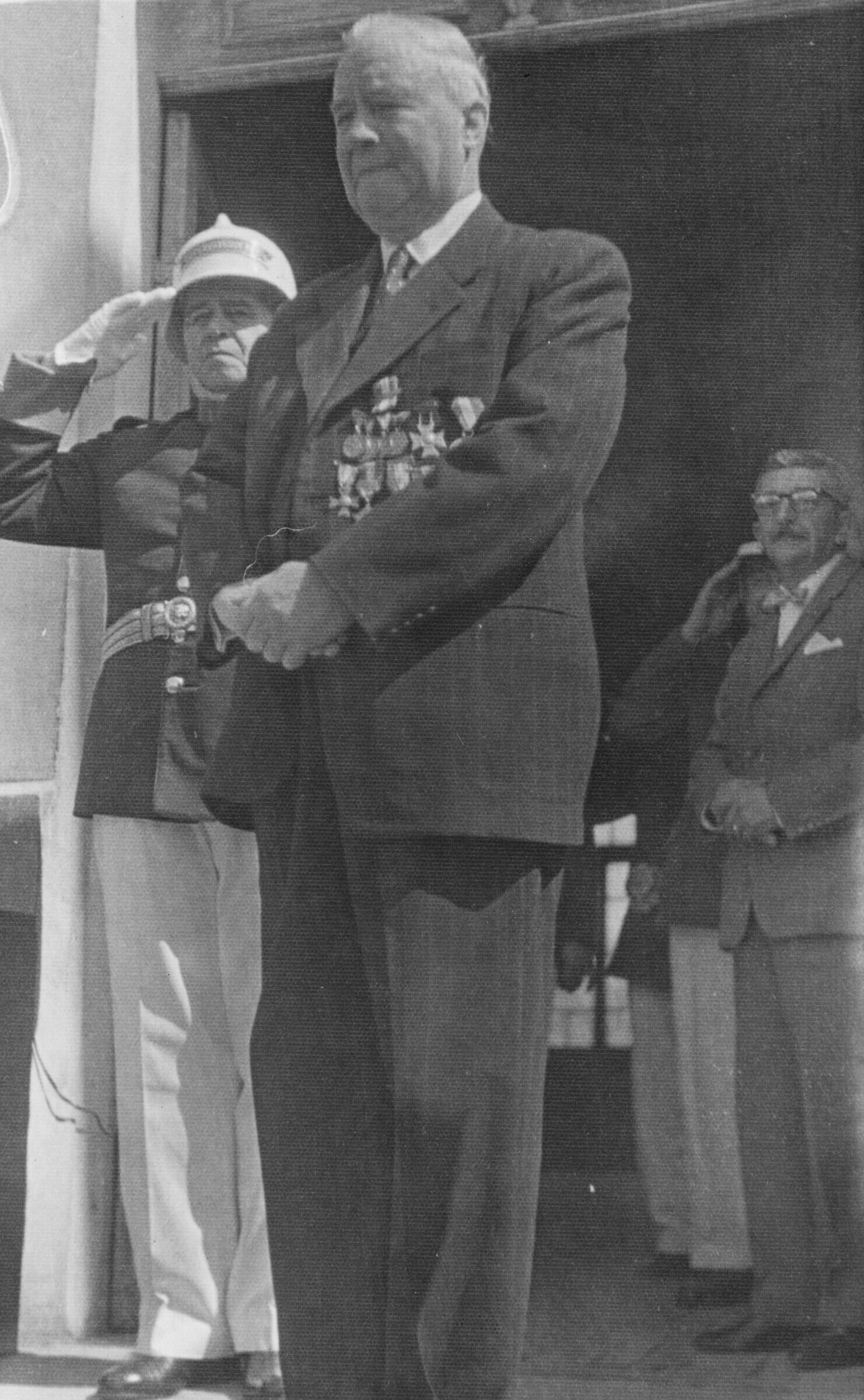 Plano entero Guillermo Gantz vestido de Traje. En su chaqueta lleva puestas todas sus medallas de reconocimiento de la institución. A sus espaldas, Ricardo Siegle realiza un saludo con la mano y también un hombre mayor está parado más en el fondo. Se encuentran en el frontis de la municipalidad de La Unión durante un homenaje realizado