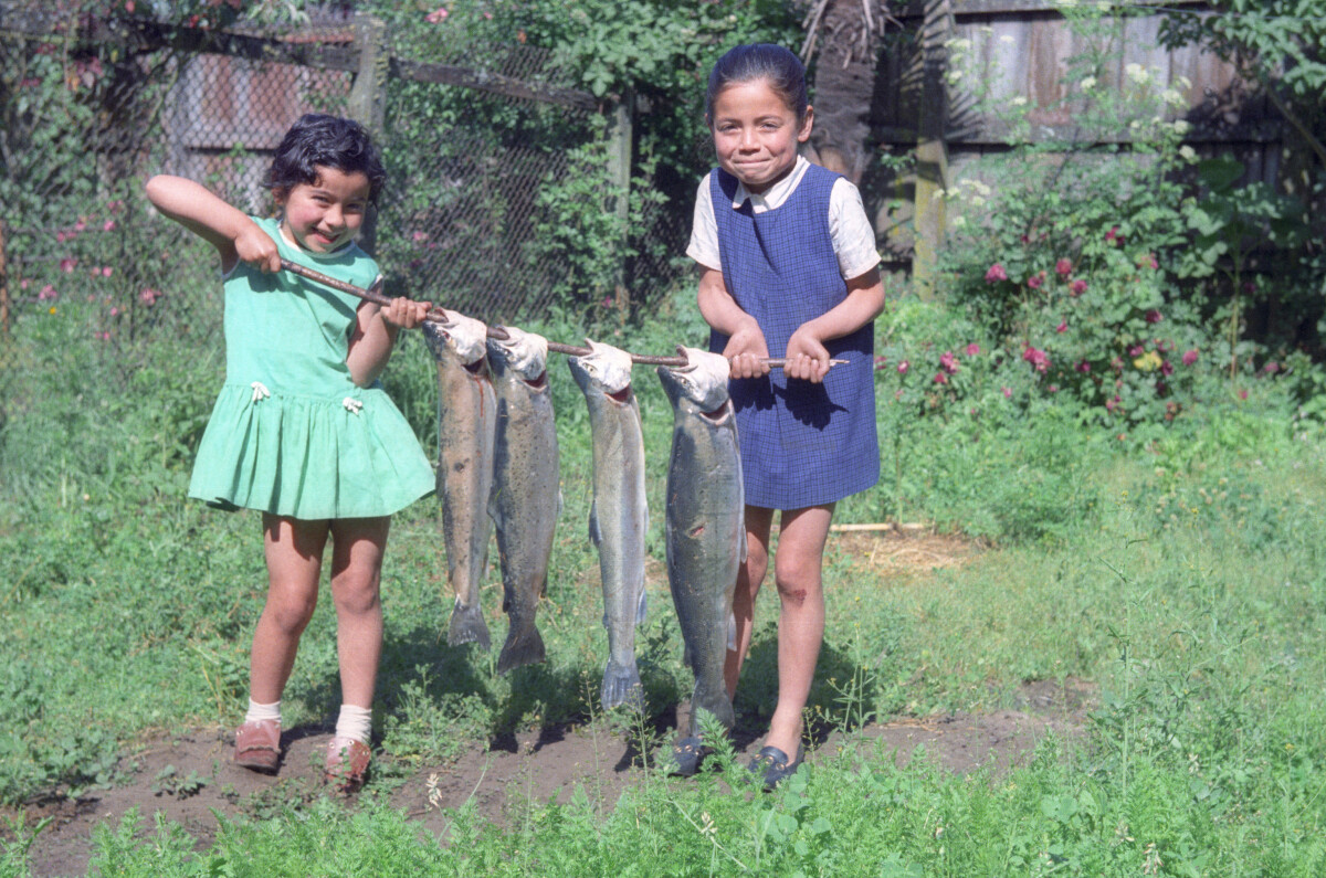 Plano entero de dos niñas sosteniendo cada extremo de un palo con cuatro pescados colgando en un patio