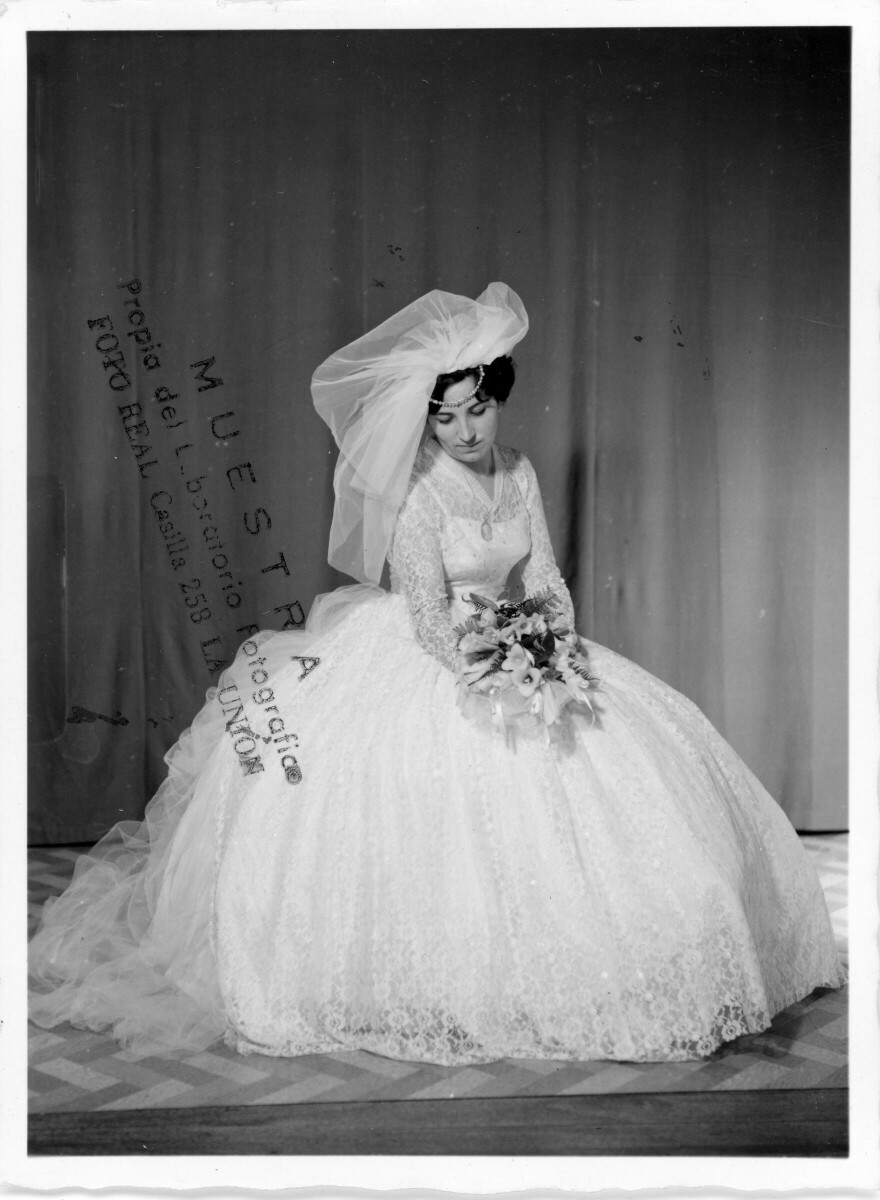 Retrato de estudio de Plano entero de Sylvia García vestida de novia, posando sentada mientras inclina su cabeza mirando el ramo de copihues blancos que tiene entre sus manos