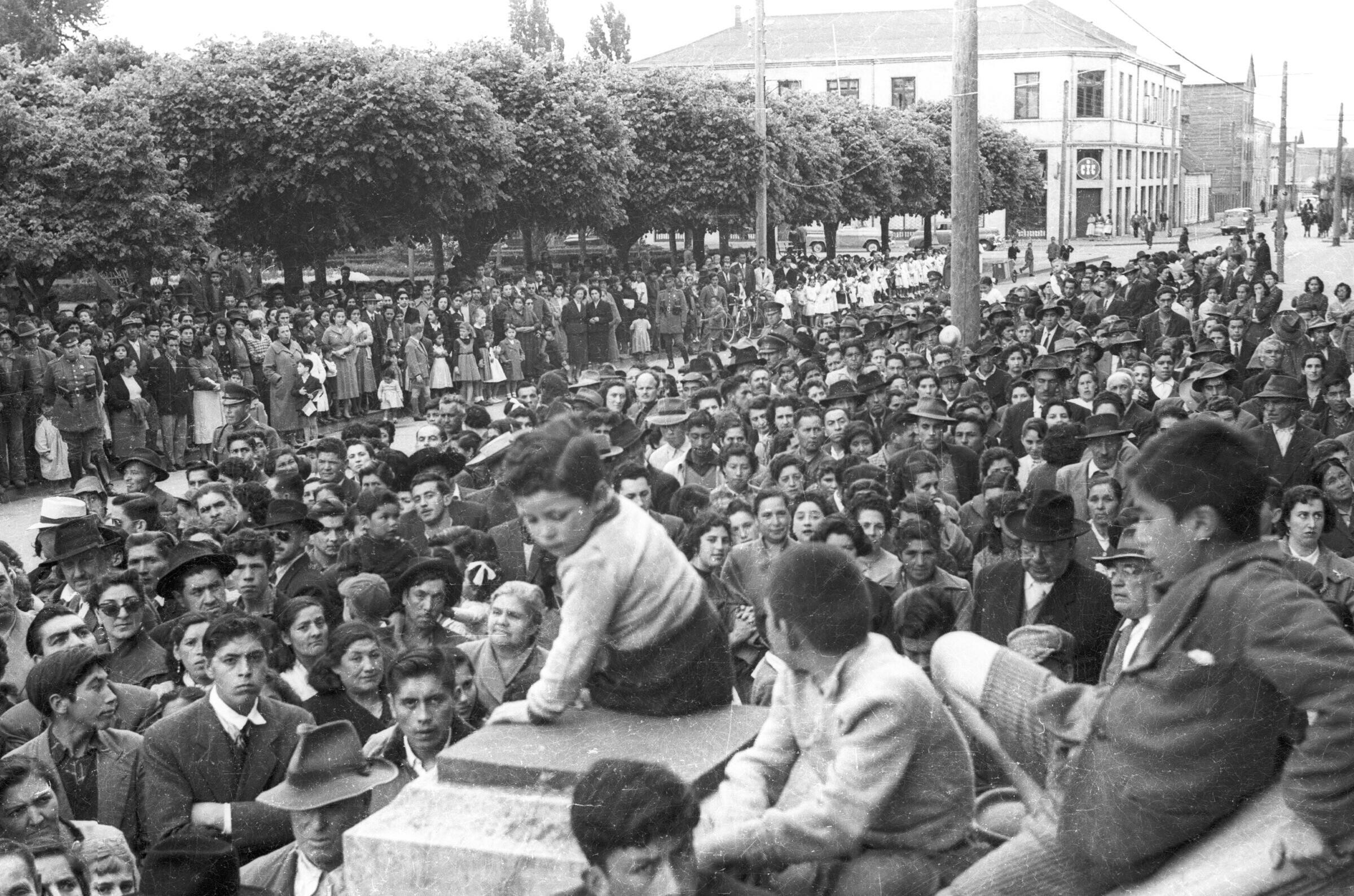 plano general de cuatro niños sentados en una estructura de cemento mientras que en el fondo, un grupo numeroso de Mujeres y Hombres se encuentran sobre la calle, durante un discurso de Carlos Vogel, realizado en el frontis de la municipalidad
