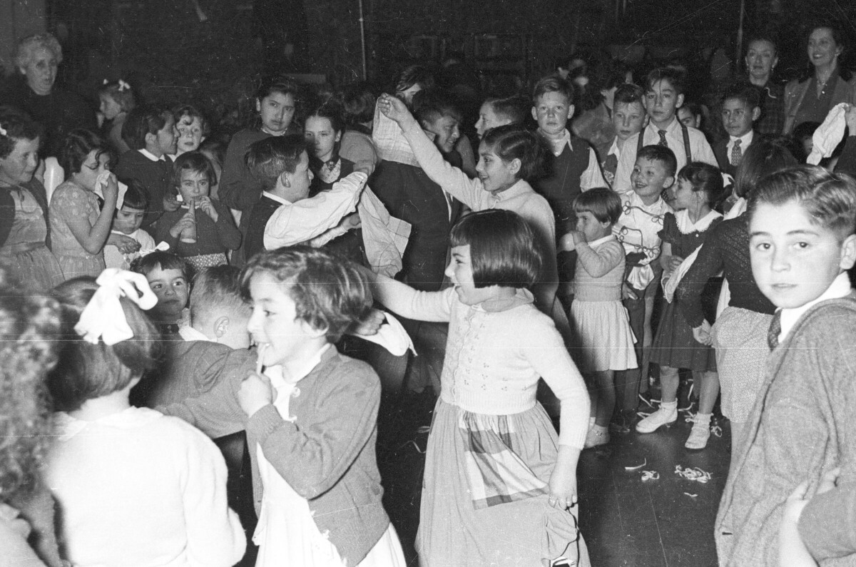 plano general de un grupo Niños bailando cueca en pareja. A su alrededor hay un grupo numeroso de Niños, Niñas y tres mujeres observándolos. Están en el interior de un local no identificado durante la celebración de fiestas patrias.