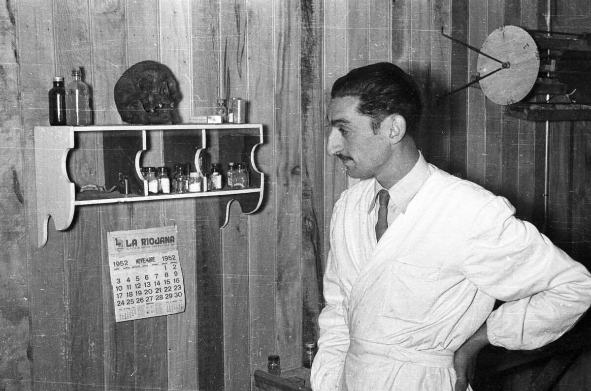 Plano entero de un hombre con bigote, vestido con un delantal blanco mirando una repisa en la pared con varios frascos y una calavera encima. El hombre tiene la mano apoyada en la zona lumbar de su espalda. Bajo la repisa hay un calendario que dice La Riojana, Noviembre 1952'