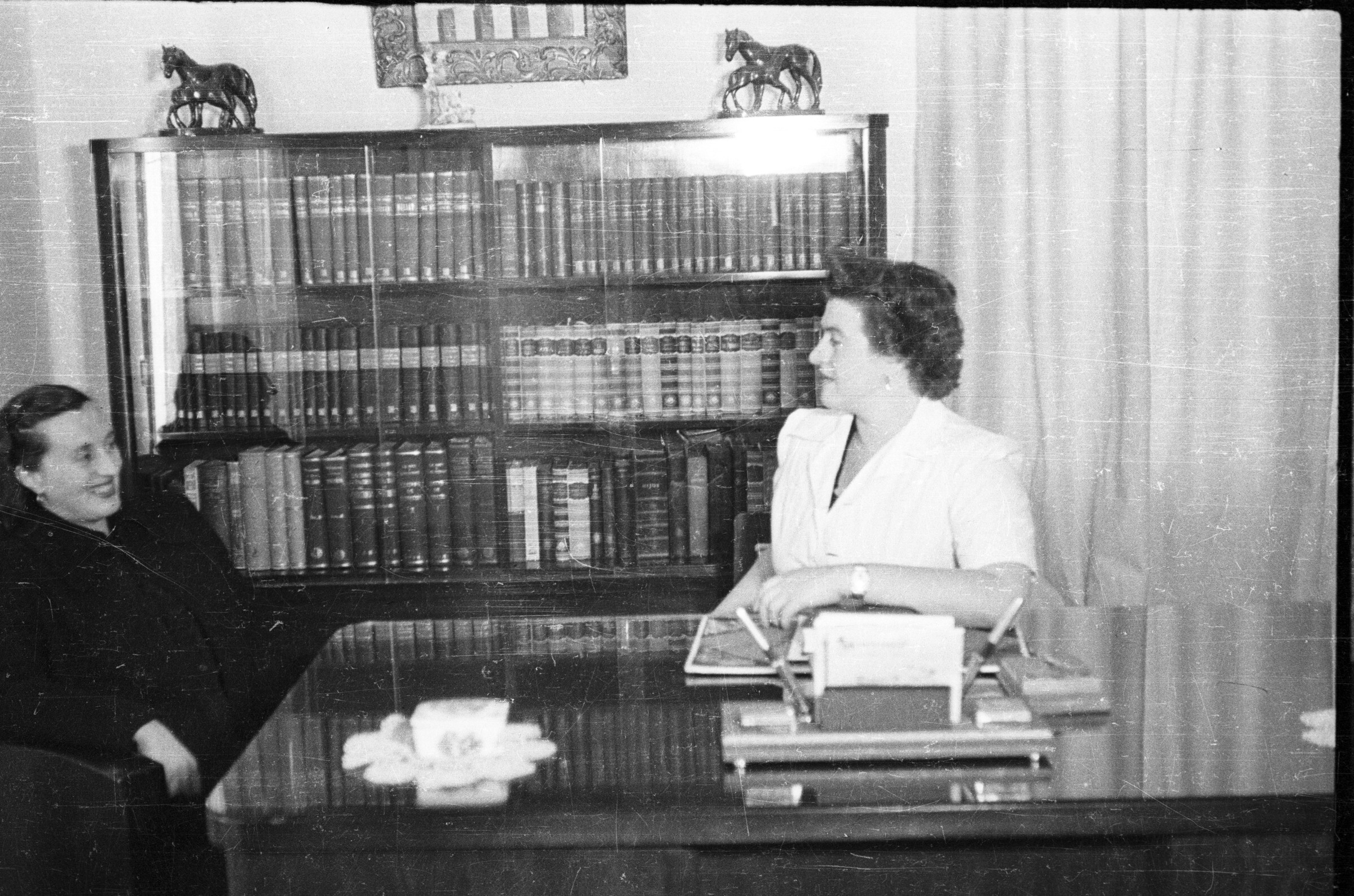 Retrato de plano entero de Guacolda y Marina Delgado, conversando en un escritorio, detrás de ellas hay un estante con libros