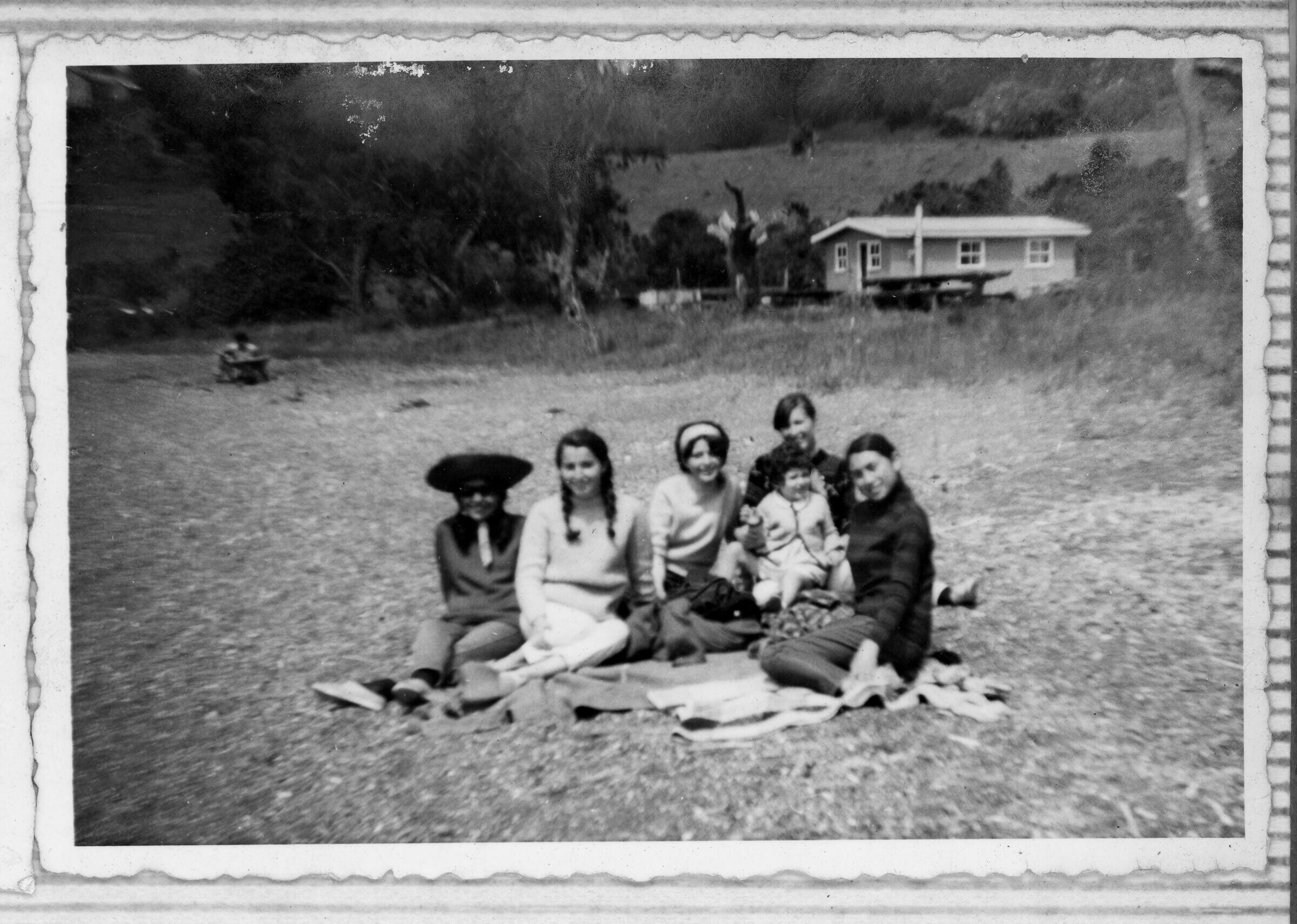 Retrato grupal de seis personas sentadas en el pasto a las afueras de una casa de campo. detrás del grupo, la casa de campo, árboles y montes.