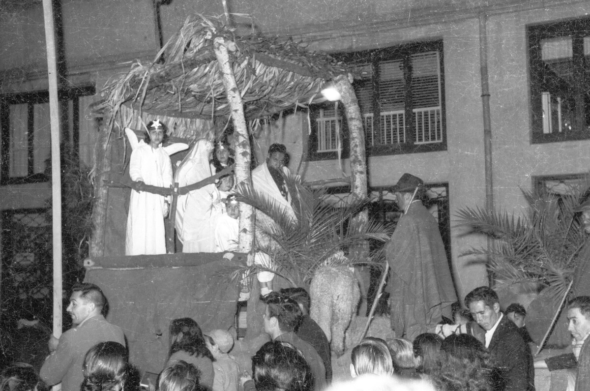 plano general de un grupo de niñas y un hombre reunidos en el interior de un carro alegórico decorado como un pesebre. A los costados Hombres y Mujeres caminando acompañándolo. Se encuentran en las afueras de un edificio de La Unión