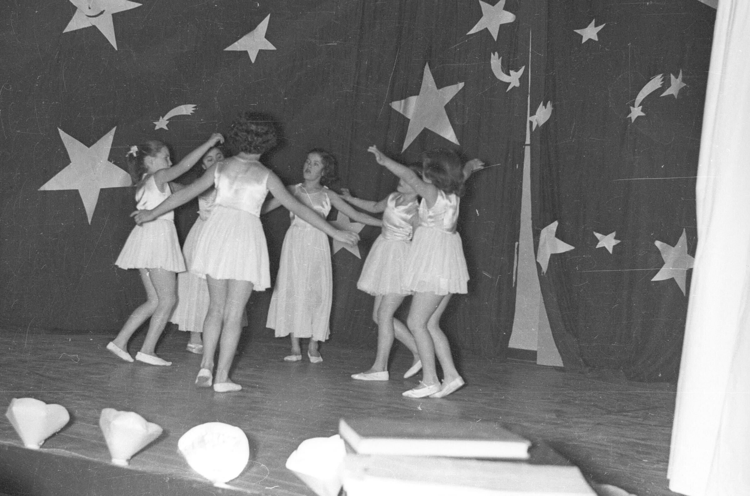 Plano entero un grupo de Niñas bailando ballet formadas en un círculo sobre el escenario. El telón del fondo está decorado con estrellas de utilerías. Están en el interior de una escuela durante una jornada de presentaciones artísticas.