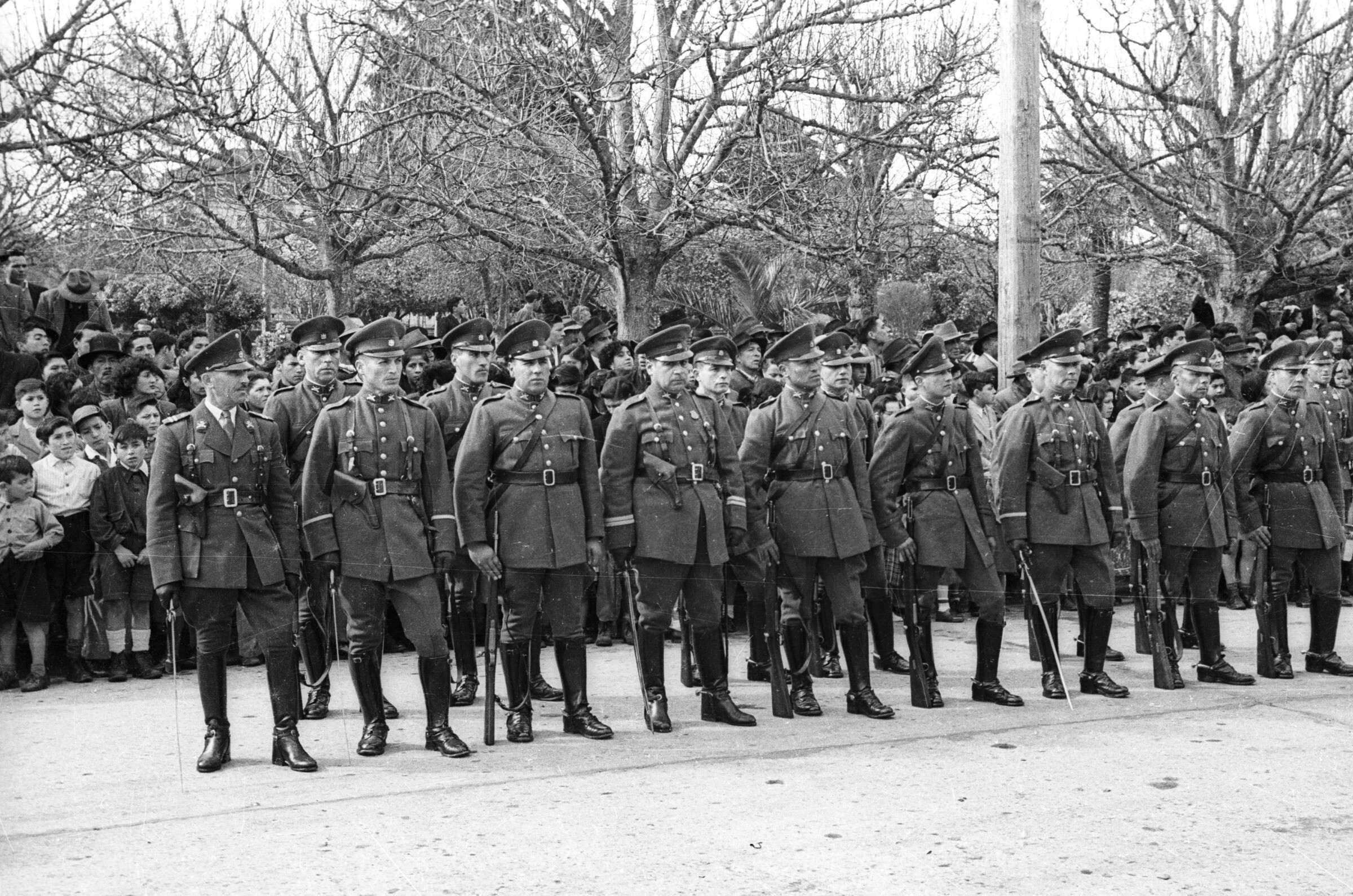 Plano entero de un grupo de carabineros formados en dos hileras durante un desfile. Llevan puesto su uniforme y algunos de ellos llevan desfundada su espada y otros afirman sus escopetas junto a su piernas. Sobre la vereda, detrás de ellos hay un grupo numeroso de Niños, Niñas, Hombres y Mujer parados, observando el desfile realizado a un costado de la plaza de La Concordia