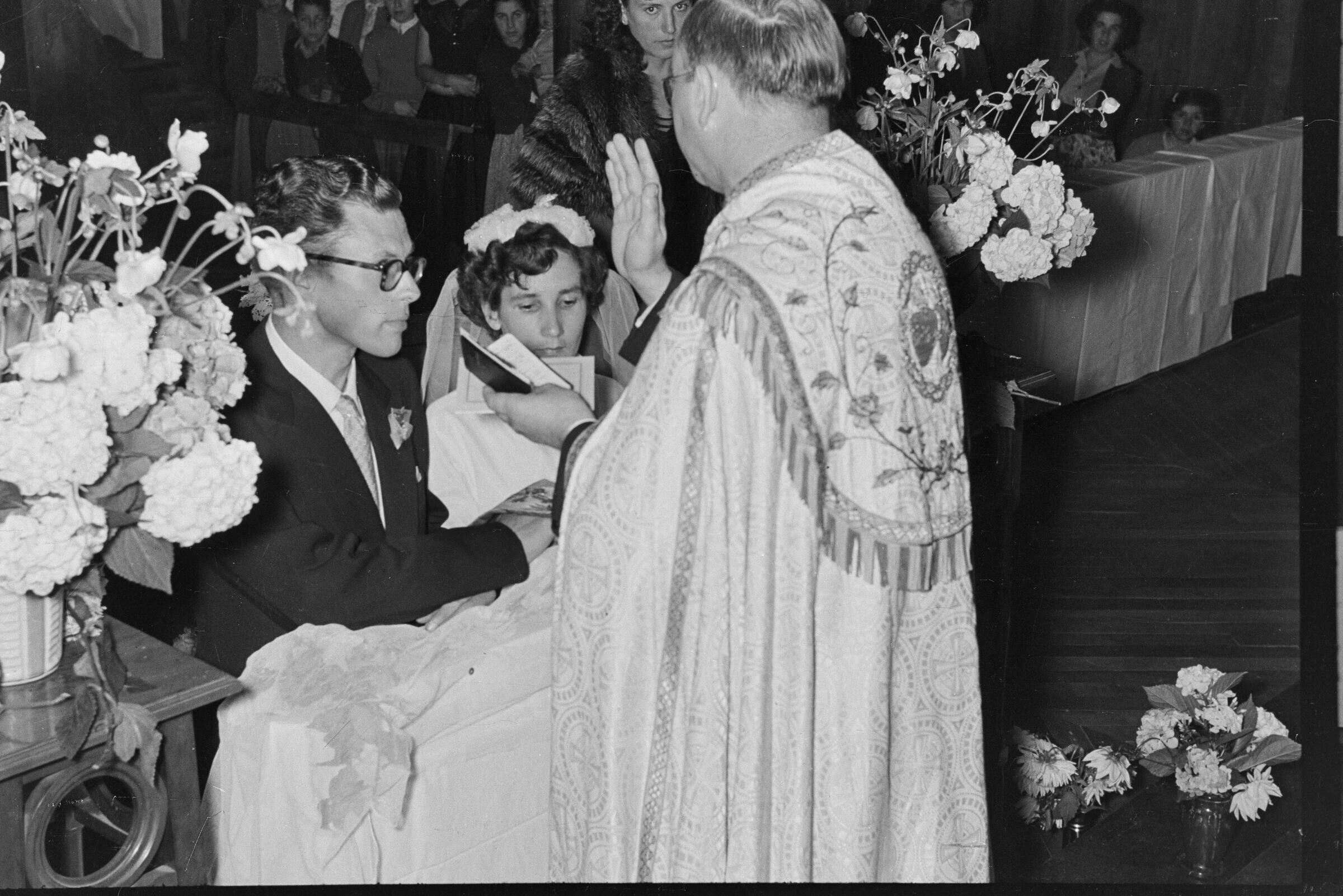 Plano entero de un sacerdote, bendiciendo a una pareja de novios, quienes se encuentran hincados frente a él, en el altar. Se encuentran en el interior de la parroquia San José lugar donde se está llevando a cabo la misa de celebración de su casamiento