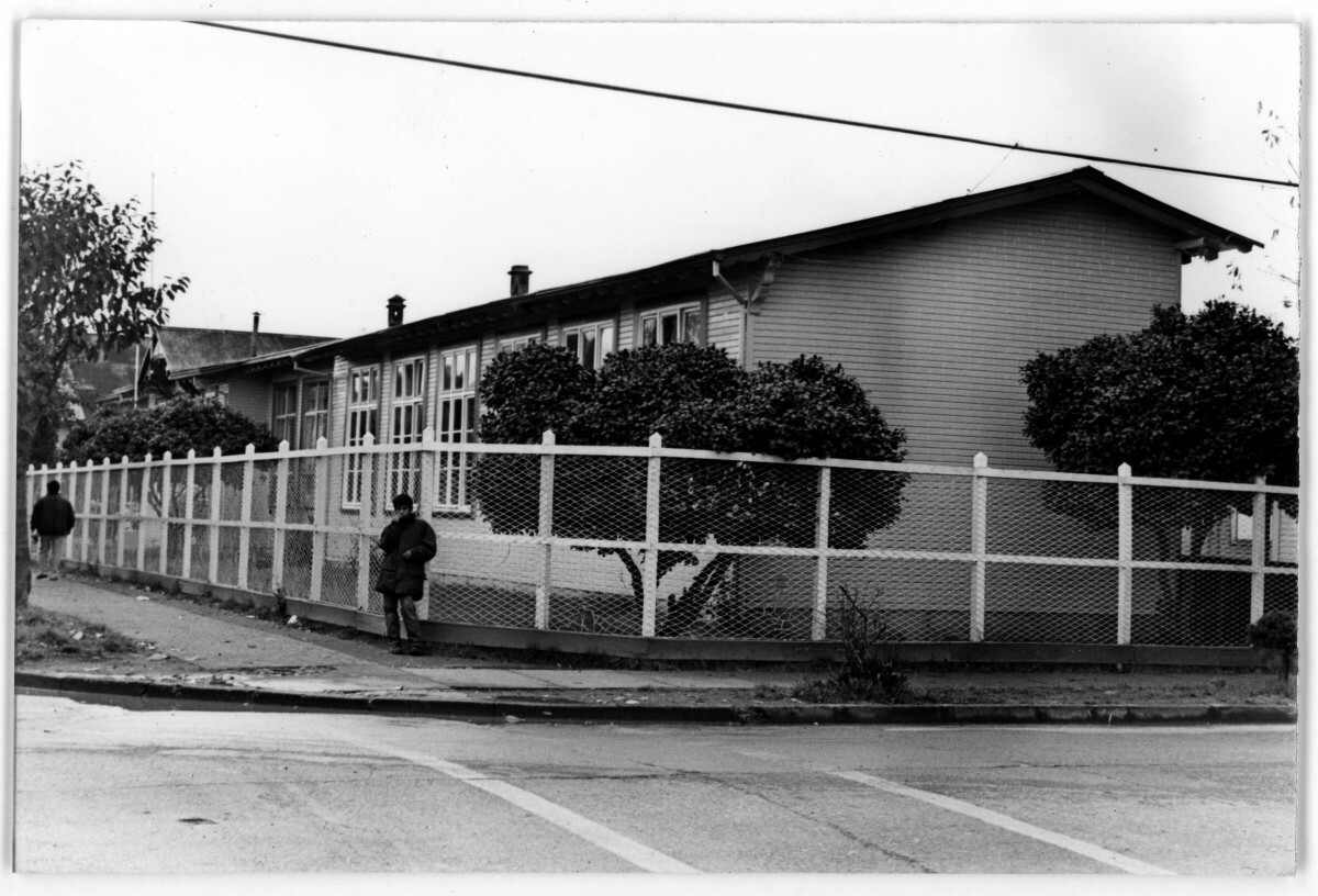 plano general de un niño apoyado en una reja mientras mira hacia la cámara. Se encuentra parado en la esquina de una cuadra, en el frontis de la Escuela Nº1 de La Unión.