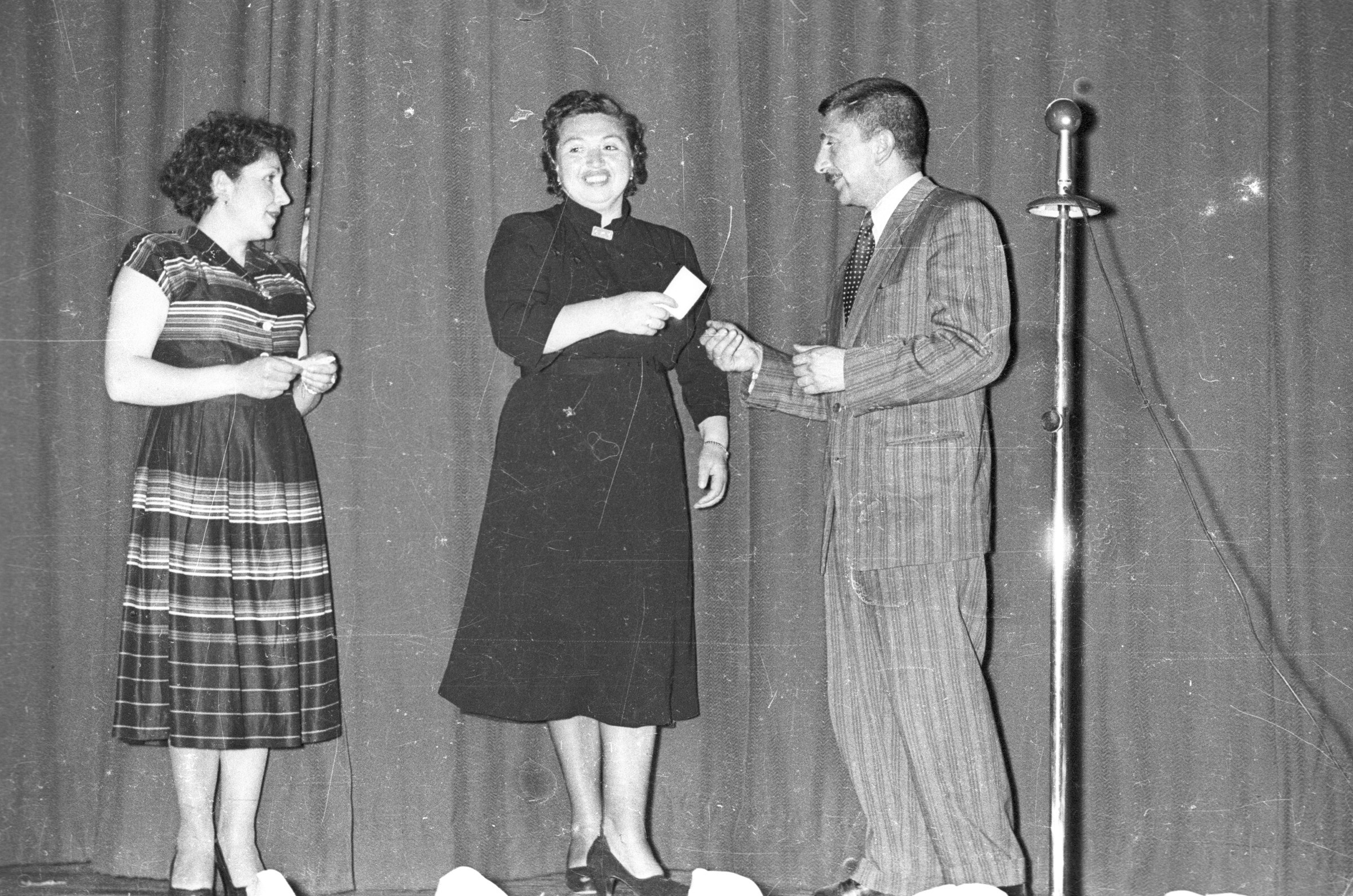 Plano entero de dos mujeres y un hombre conversando parados sobre el escenario durante una actividad escolar en el interior del gimnasio de la Escuela superior de Niñas Nº2