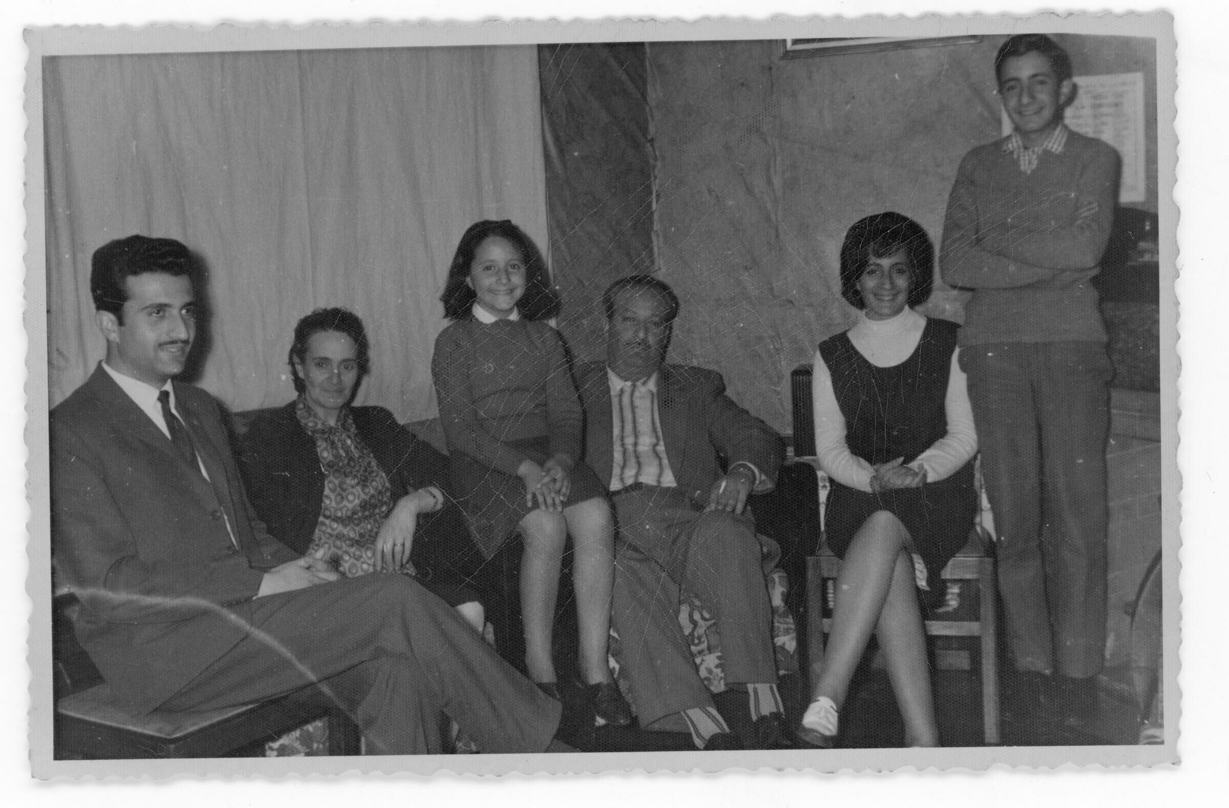 Retrato grupal de plano entero de dos hombres, dos mujeres, una niña y hombre joven, posando sentados a excepción del ultimo mencionado. Se encuentran en el interior de una casa