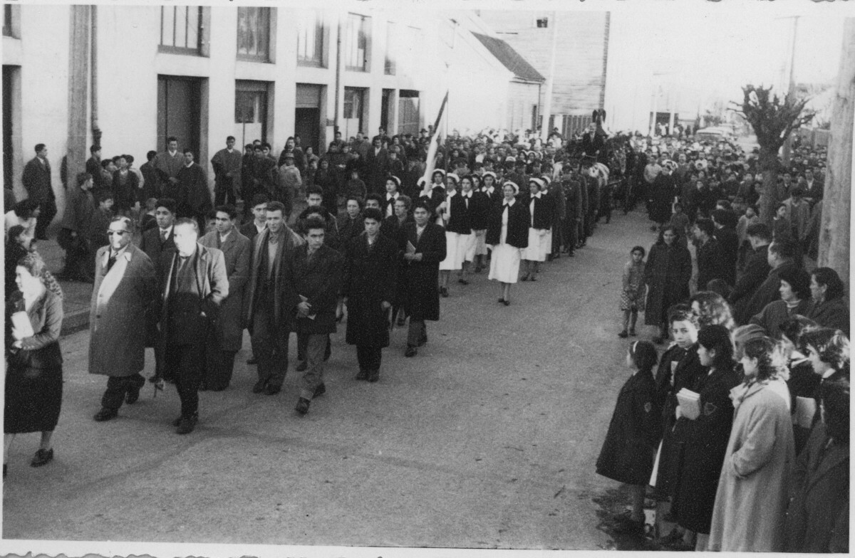plano general de un cortejo fúnebre. Un grupo de hombres, mujeres de la cruz roja y carabineros, lideran la caravana. A sus espaldas avanza la carroza y el resto de las personas que se dirigen al cementerio. A cada costado de la calle, hay otro grupo numeroso de personas que están paradas observándolos transitar.