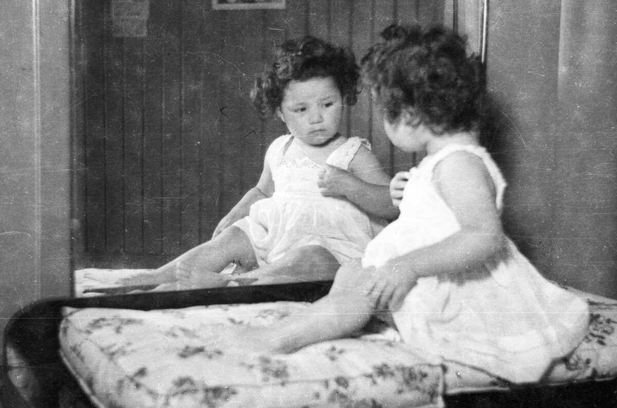 Plano entero de un niña sentada frente a un espejo, observando su reflejo.