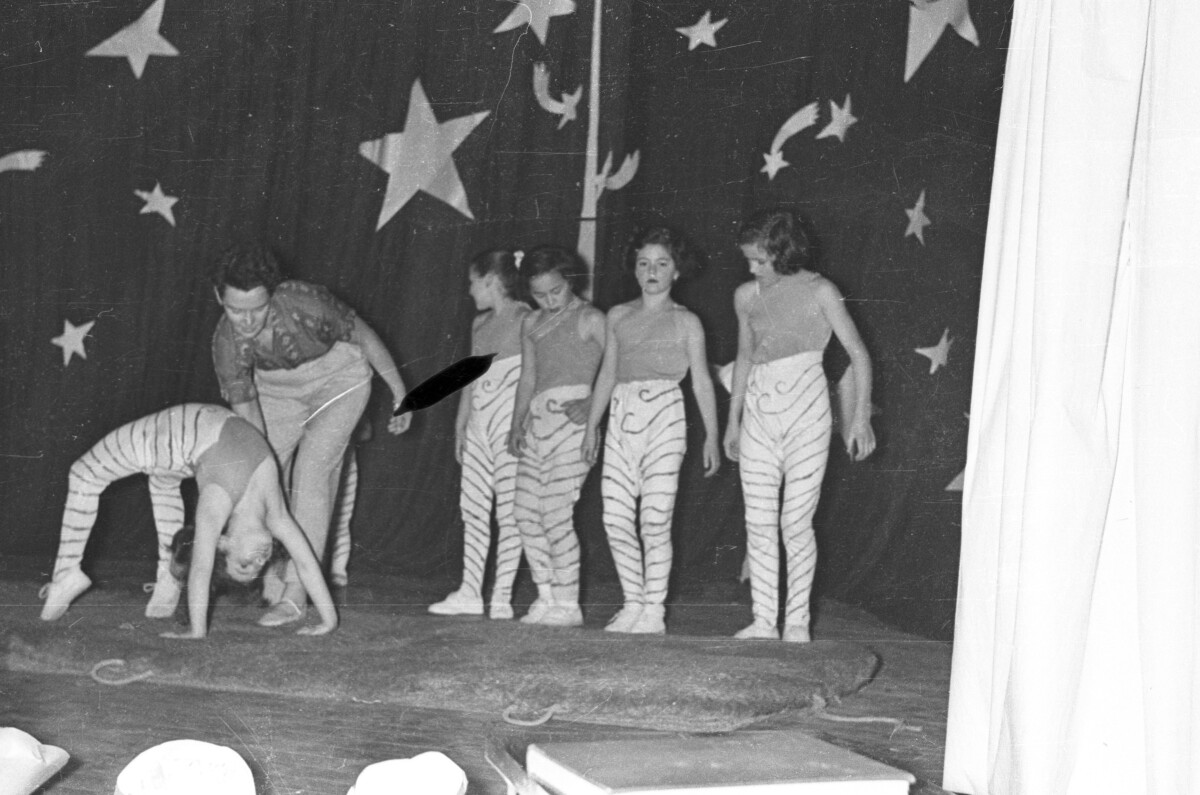 Plano entero de una mujer tomando por la espalda a una Niña que está realizando la posición del arco invertido sobre el escenario. Junto a ellas hay un grupo de Niñas formadas en una hilera mientras realizan una presentación de ballet en el escenario de una escuela durante una jornada de presentaciones artísticas