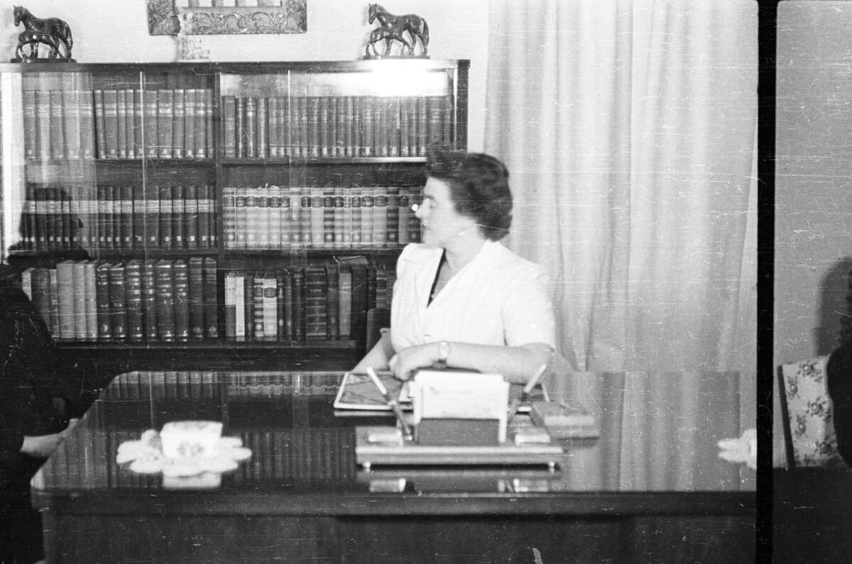 Retrato de plano entero de dos mujeres sentadas conversando en escritorio, detrás de ellas hay un estante con libros