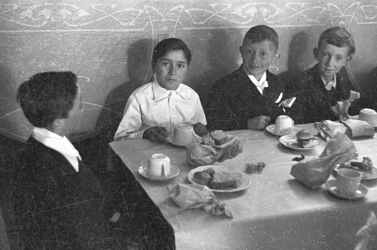 Plano entero de cuatro niños sentados alrededor de una mesa sobre la cual hay platos con galletas y queques. Tres de ellos están mirando hacia la cámara mientras que el cuarto está sentado de perfil sobre su silla. Están en el interior de una infraestructura no identificada celebrando su primera comunión