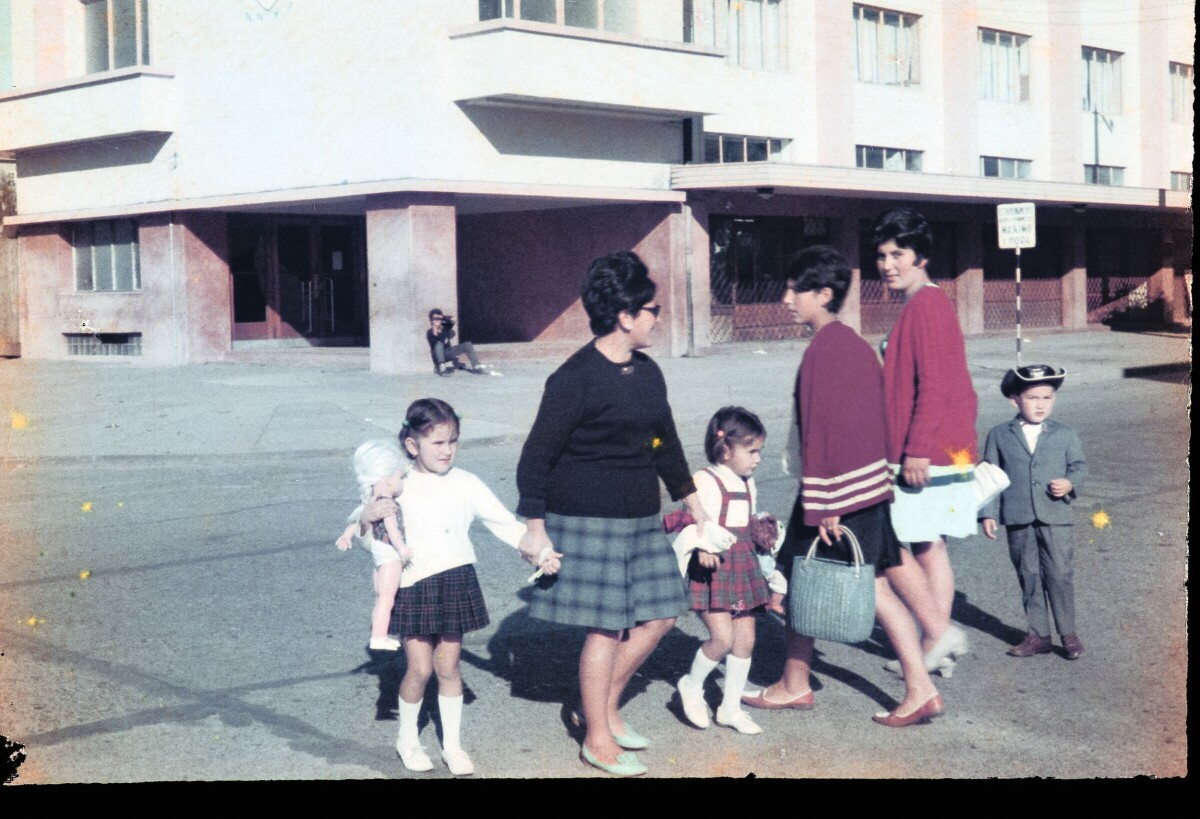 Retrato de plano entero de Mujer con Vestido caminando junto a dos niñas y un niño mientras dos mujeres pasan cerca de ellos. Frente a ellos la municipalidad de río Bueno
