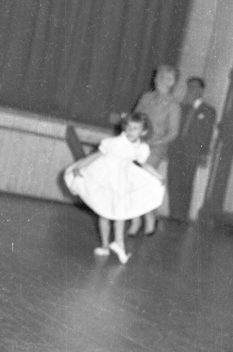 Plano entero de una Niña con Vestido, levantando la punta de su falda durante una presentación artística realizada en el interior de una estructura no identificada. fotografía borrosa