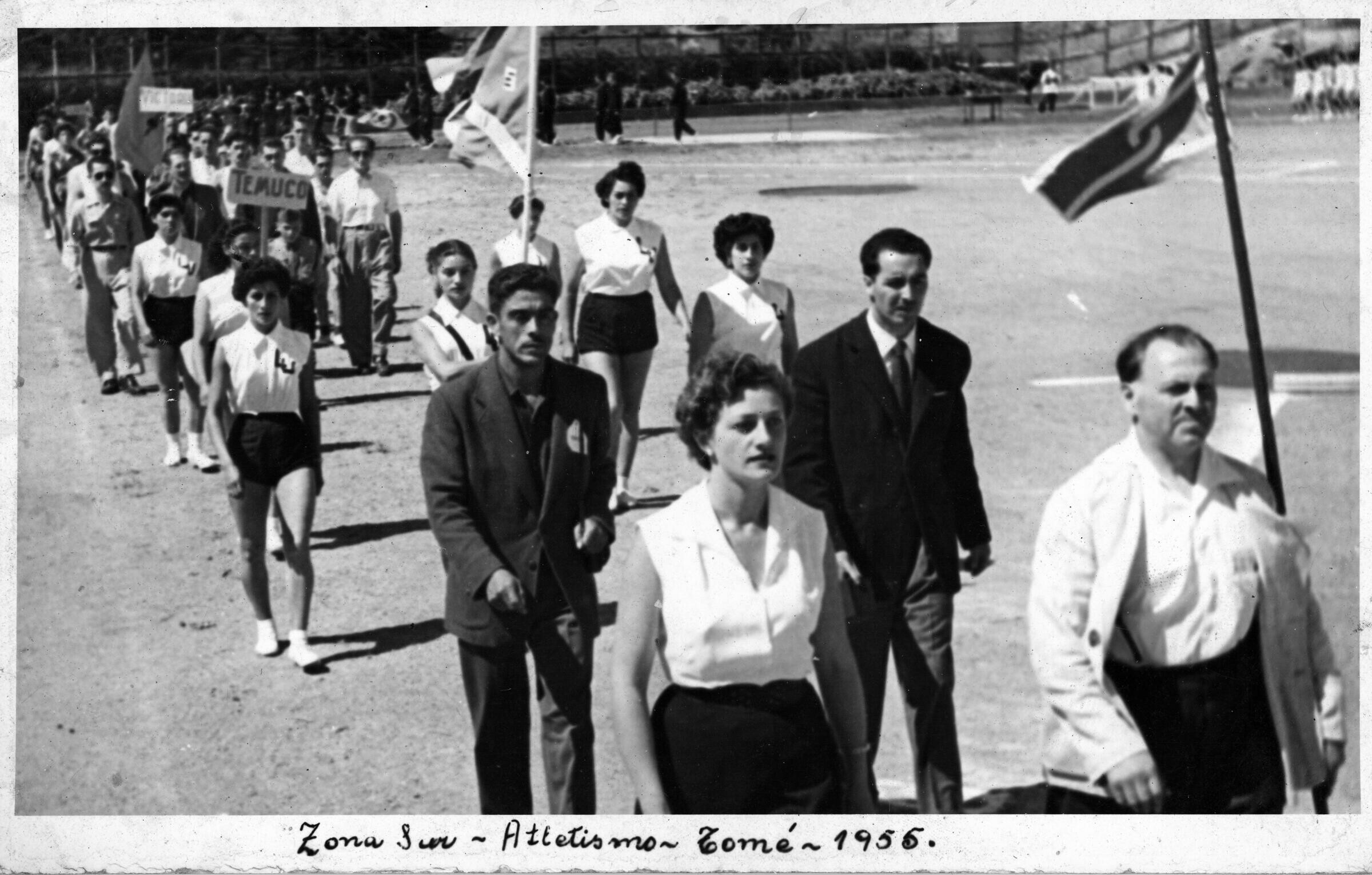 -plano general de un grupo de atletas y hombres desfilando en una hilera. Algunos llevan consigo banderas y en el medio alguien lleva un letrero que dice --Temuco--. Todas las mujeres llevan en sus poleras las iniciales --LU-- bordadas en el pecho. En el frente, la primera mujer es Xenia Vogel. Raquel Latorre está caminando al lado izquierdo de la mujer que sostiene una bandera y detrás de ellas, al costado derecho, la mujer de mayor estatura es Pradelia Delgado. Se encuentran en un zonal sur de atletismo realizado en Tomé.-