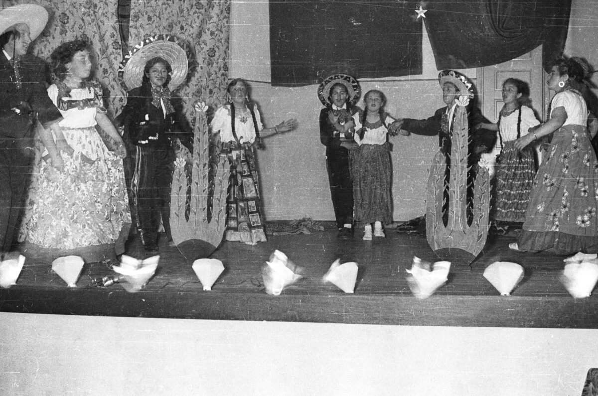 plano general de un grupo de Niñas y Niños disfrazados. Los Niños visten como charros mexicanos y las Niñas visten como las conocidas chinas poblanas de México. Están ubicados en semicírculo alrededor de dos cactus de utilería mientras presentan una obra de teatro sobre el escenario de su escuela.