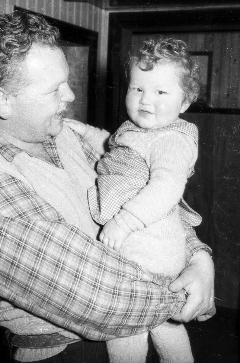 Retrato de plano medio de Hombre con camisa y chaleco sin mangas con Bebé en brazos, ambos sonriendo