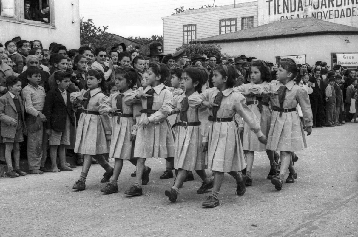 Plano entero de un grupo de niñas, pertenecientes a una brigada no identificada, desfilando formadas en dos hileras mientras realizan un saludo al pecho. En un costado un grupo numeroso de personas las observa avanzar desde el frontis de la municipalidad