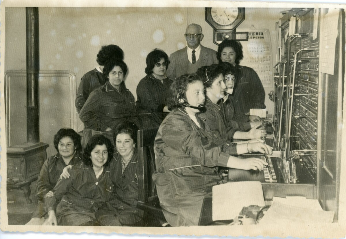 Retrato de plano entero de un grupo de trabajadoras posando por detrás de las sillas de tres operadoras telefónicas mientras éstas trabajan frente a una maquinaria no identificada. En el fondo, hay un hombre de Traje, parado entre otro grupo de mujeres. Lugar no identificado