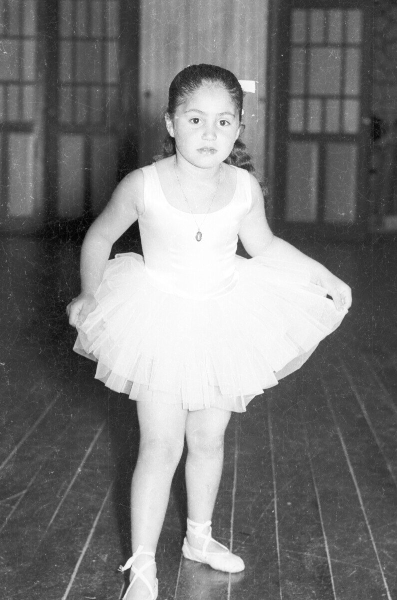 Retrato de plano entero de una niña posando de frente. Está caracterizada como bailarina de ballet. Se encuentra en una estructura no identificada durante una velada de presentaciones artísticas