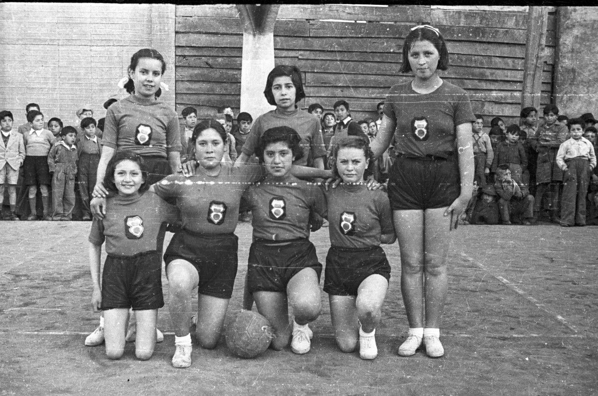 Retrato de plano entero de un equipo de baloncesto de Niñas , están posando de pie y arrodilladas junto a la pelota, en el fondo hay un grupo numeroso de Niños apoyados en la pared de la iglesia que las observan a un costado de la cancha.