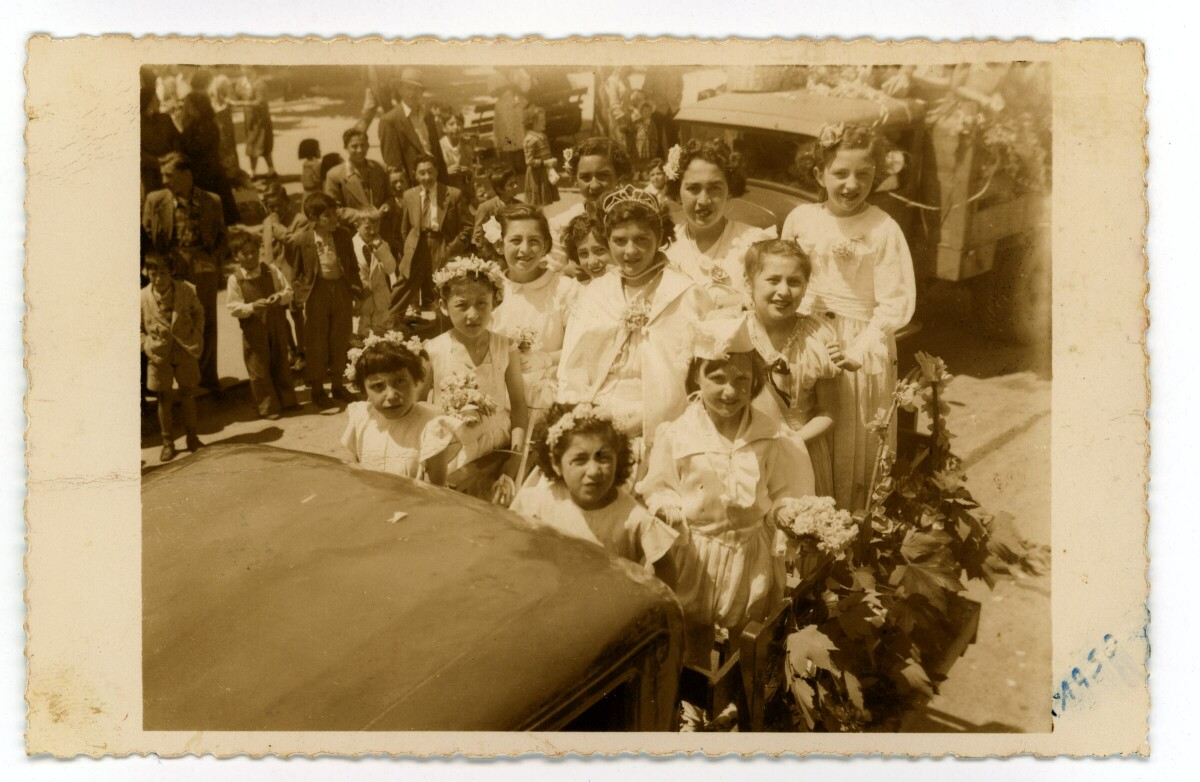 Grupo de niñas sobre caja de camioneta, todas vestidas de blanco y una de ellas vestida de reina, con una tiara. detrás, en la calle, un grupo de personas observa pasar la camioneta.