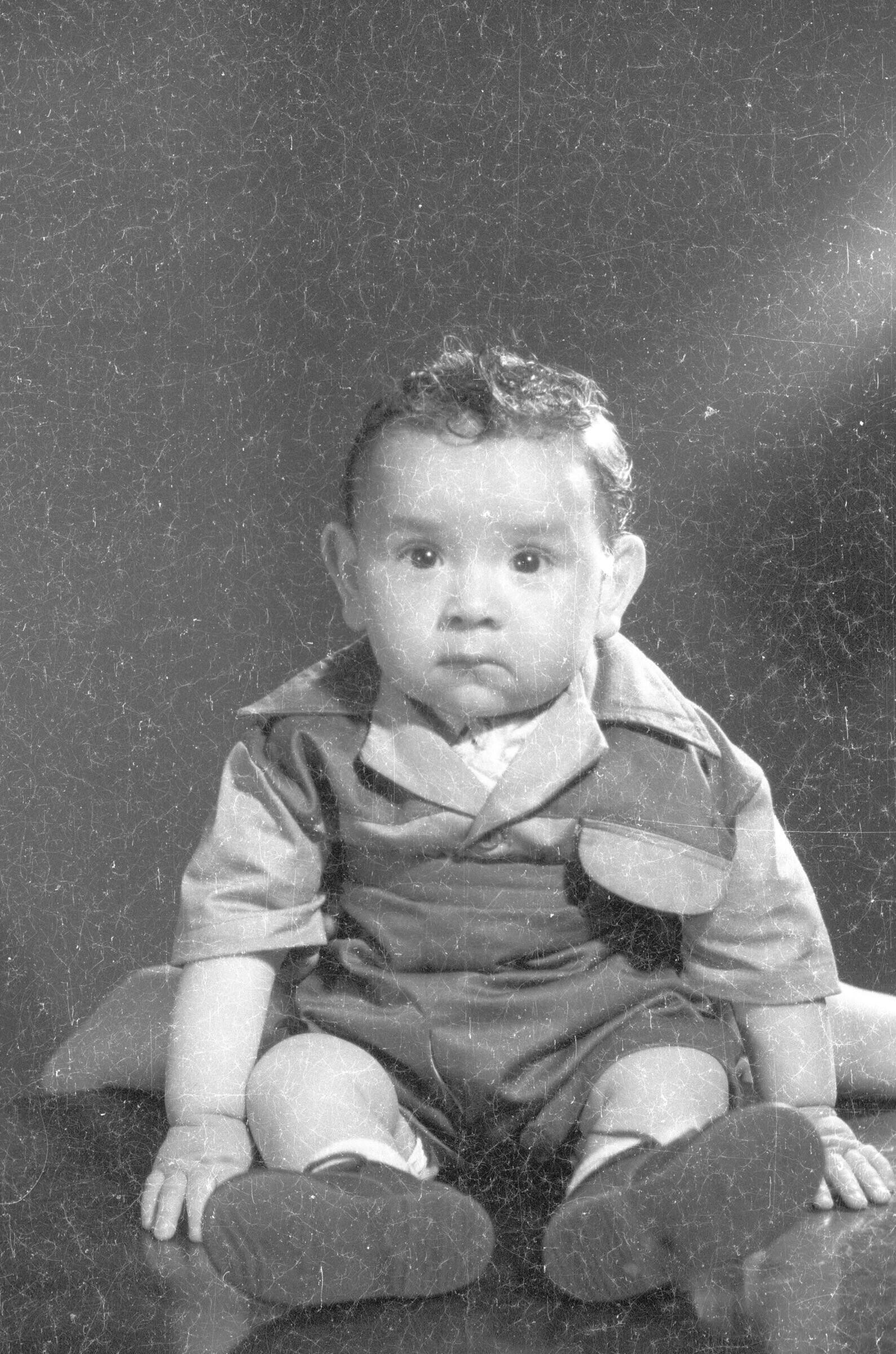 Retrato de estudio de Plano entero de un bebé posando sentado sobre una mesa, mientras que a sus espaldas hay una persona escondida afirmandolo