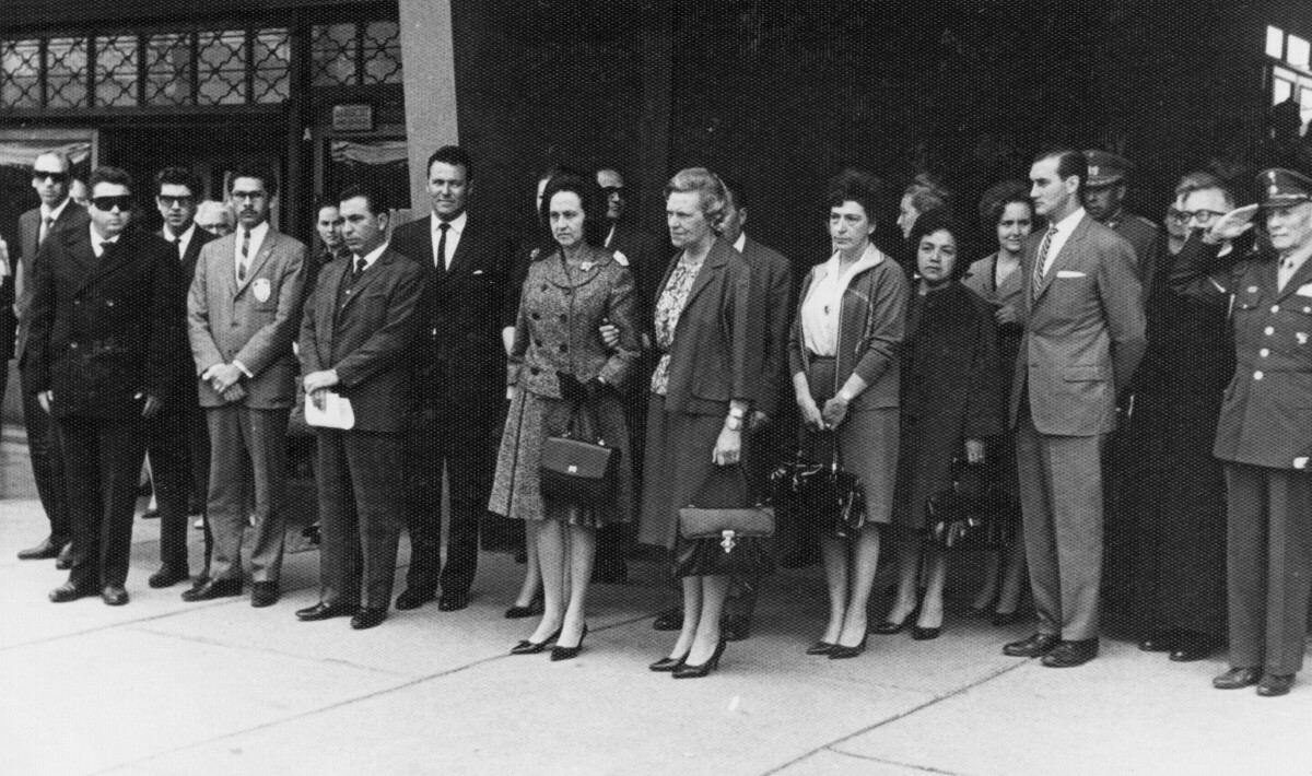 plano general de un grupo de Hombres y Mujeres parados sobre una vereda. En el medio. Olga Boettcher está tomada del brazo de Eva Sámano, primera dama de México, durante un homenaje realizado para ella en su visita a la ciudad de La Unión