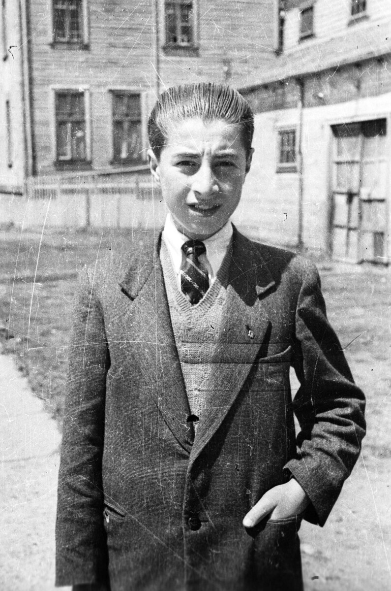Retrato de plano medio de un joven posando. Tiene una mano en el bolsillo de su traje mientras está parado en una vereda junto a la calle.