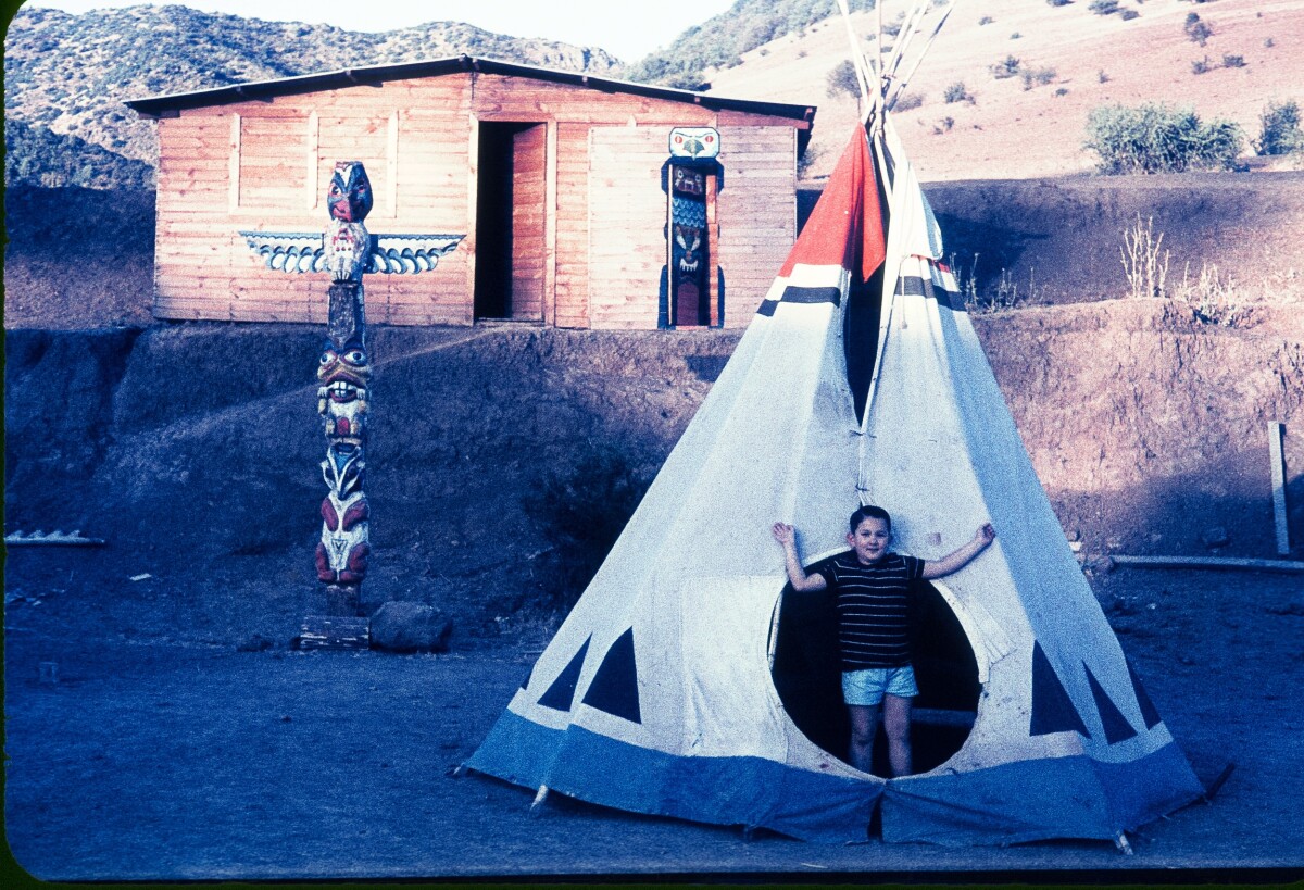 Plano entero de niño dentro de una carpa india, detrás un totem indio y una cabaña en un lugar con cerros y poca vegetación